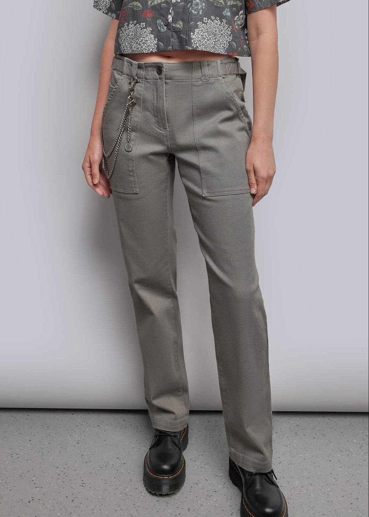 A model stands against a light gray studio background wearing a short-sleeve button-up shirt with a muted floral print in gray, green, and burgundy, paired with olive-gray trousers and black shoes.