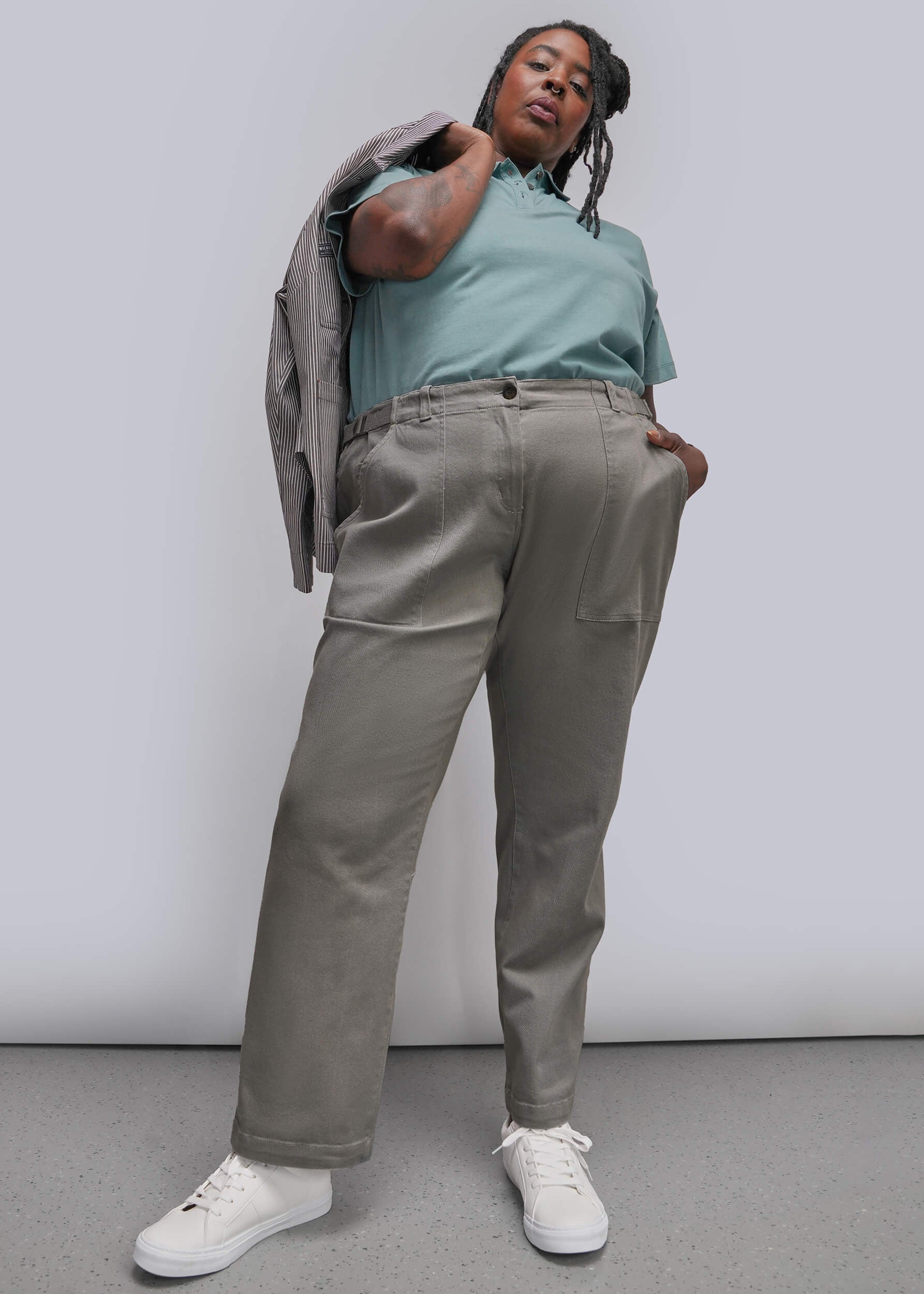 1x=A model wearing high-waisted, wide-leg taupe trousers, a teal polo shirt, and white sneakers, with a gray blazer draped over her shoulder, set against a plain light gray studio background.