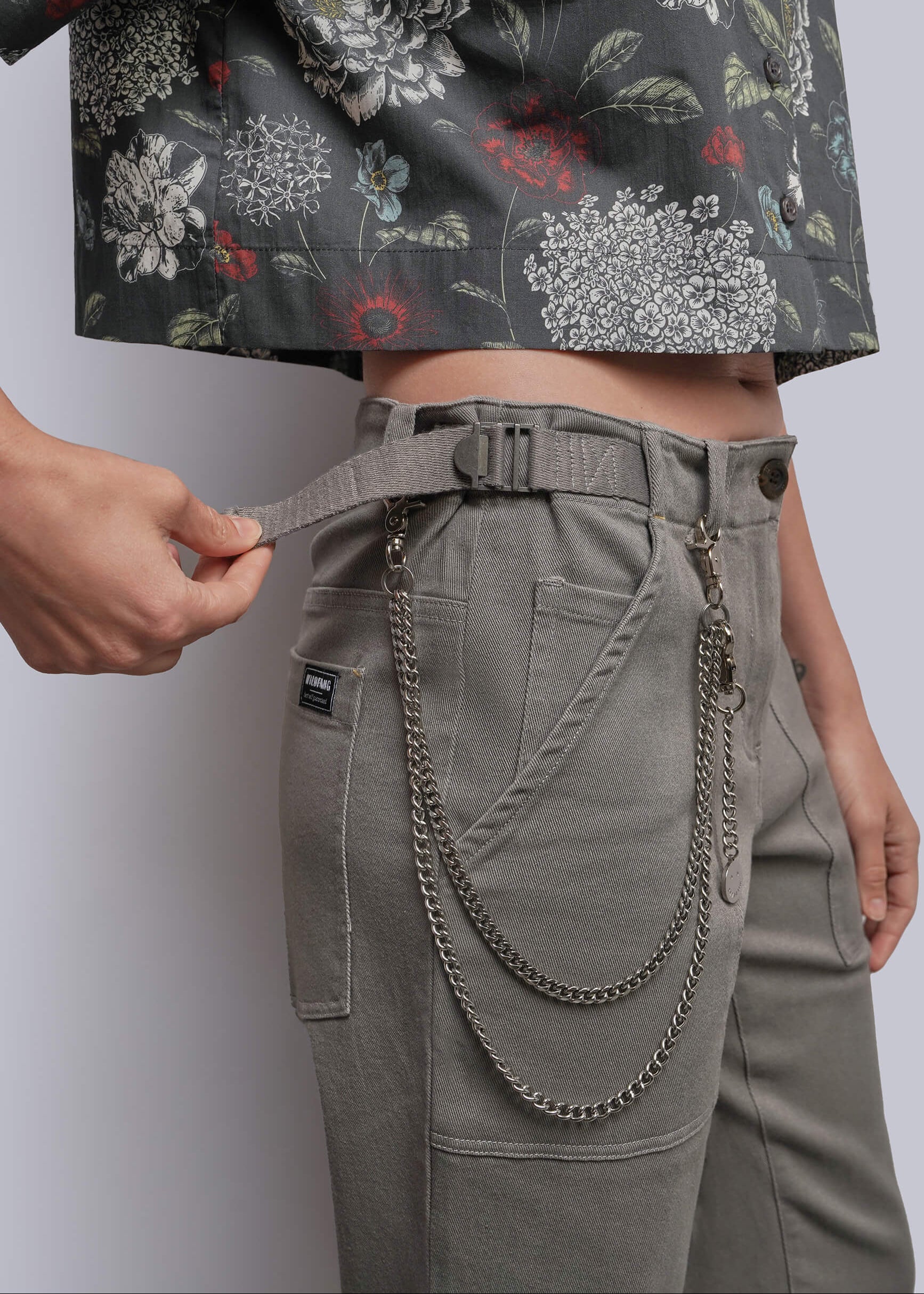 Close-up of a person wearing high-waisted gray cargo-style pants with a matching belt and hanging metal chain detail, paired with a cropped floral top against a light gray studio background.