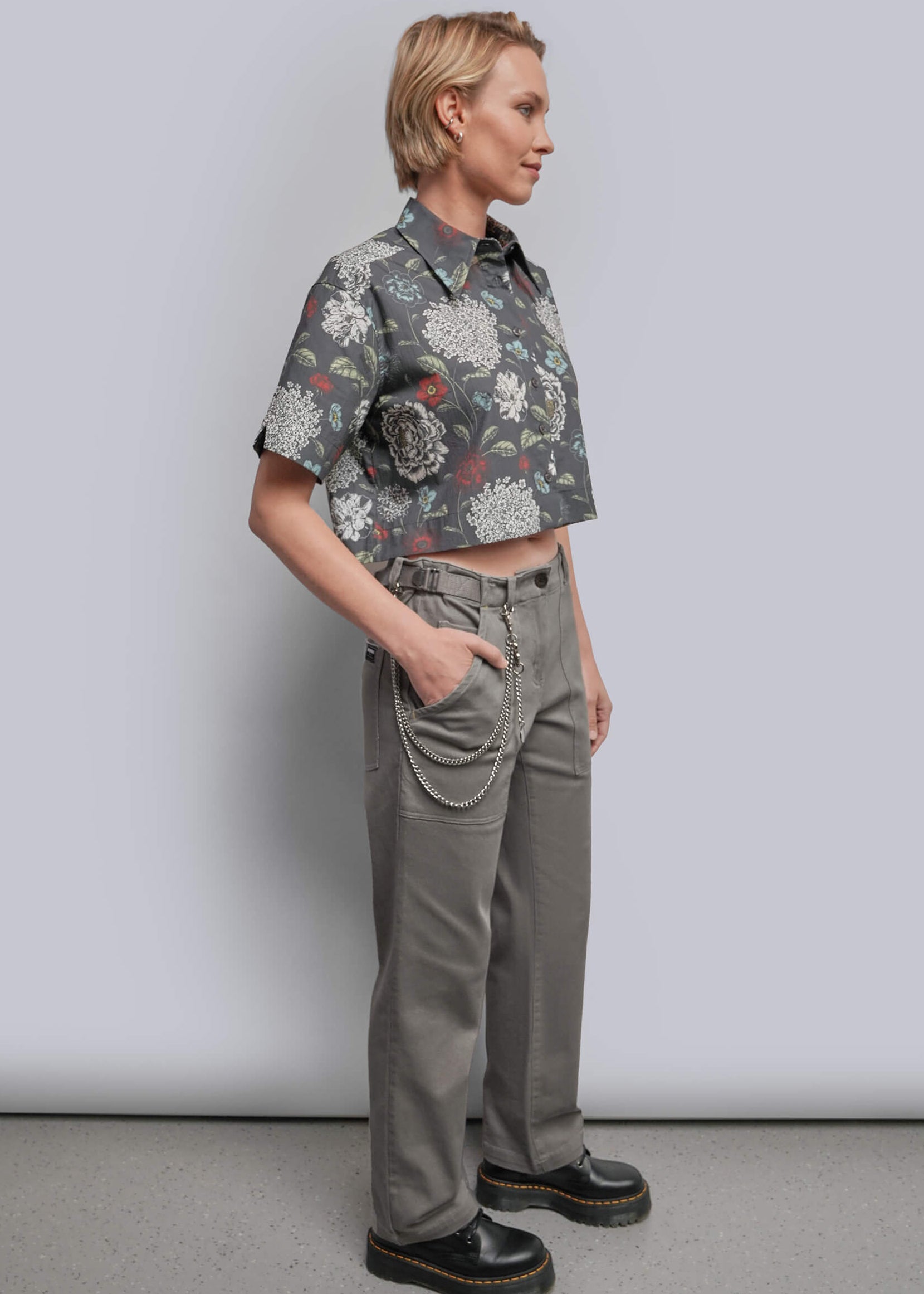 S=A model stands in profile wearing a short-sleeve button-up shirt with a muted floral print in gray, white, and burgundy, paired with gray trousers, black chain belt, and black platform shoes against a light gray studio background.