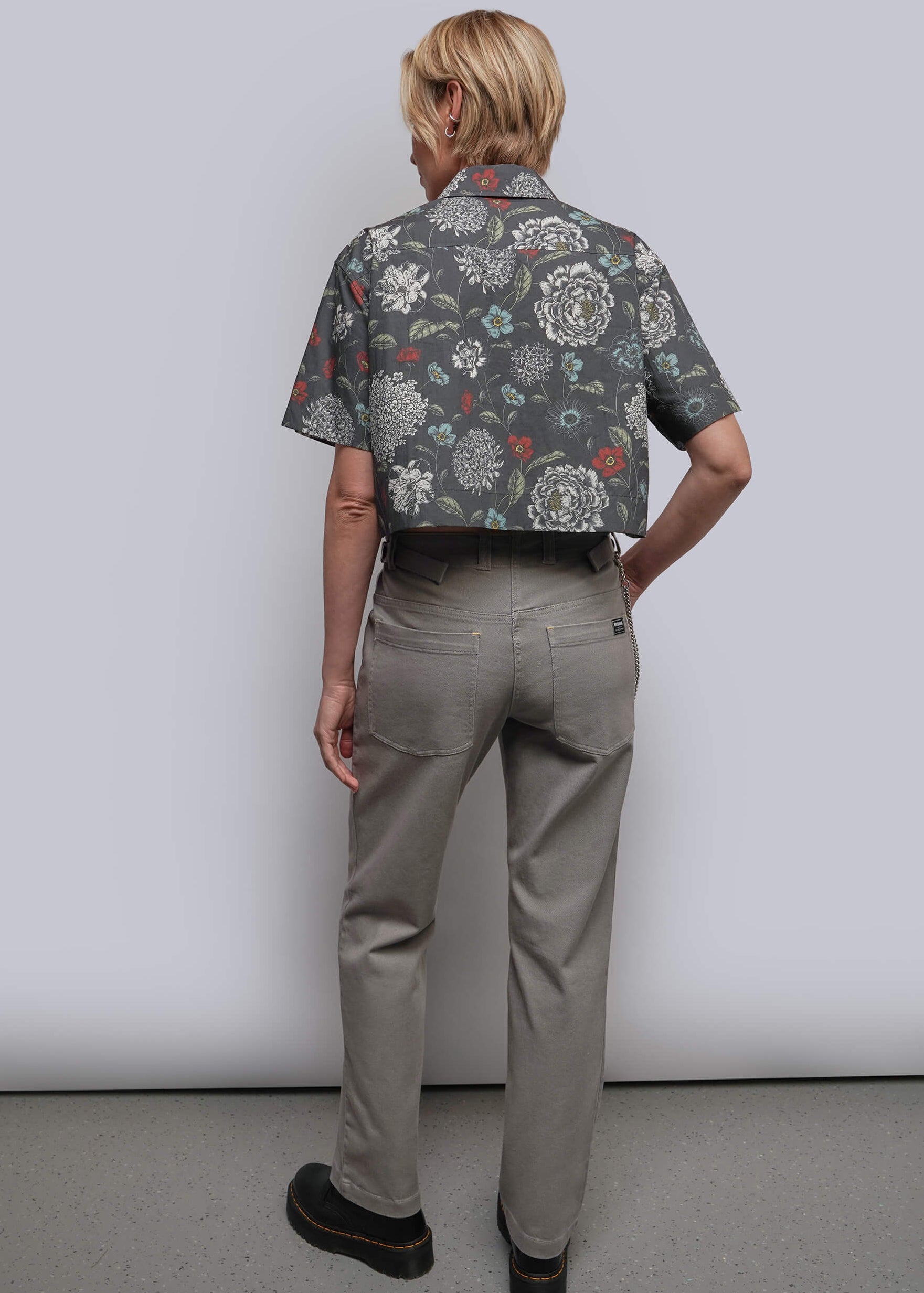 S=A model shown from the back wearing a cropped short-sleeve button-up shirt with a dark gray floral pattern and light gray trousers, standing against a plain light gray studio background.