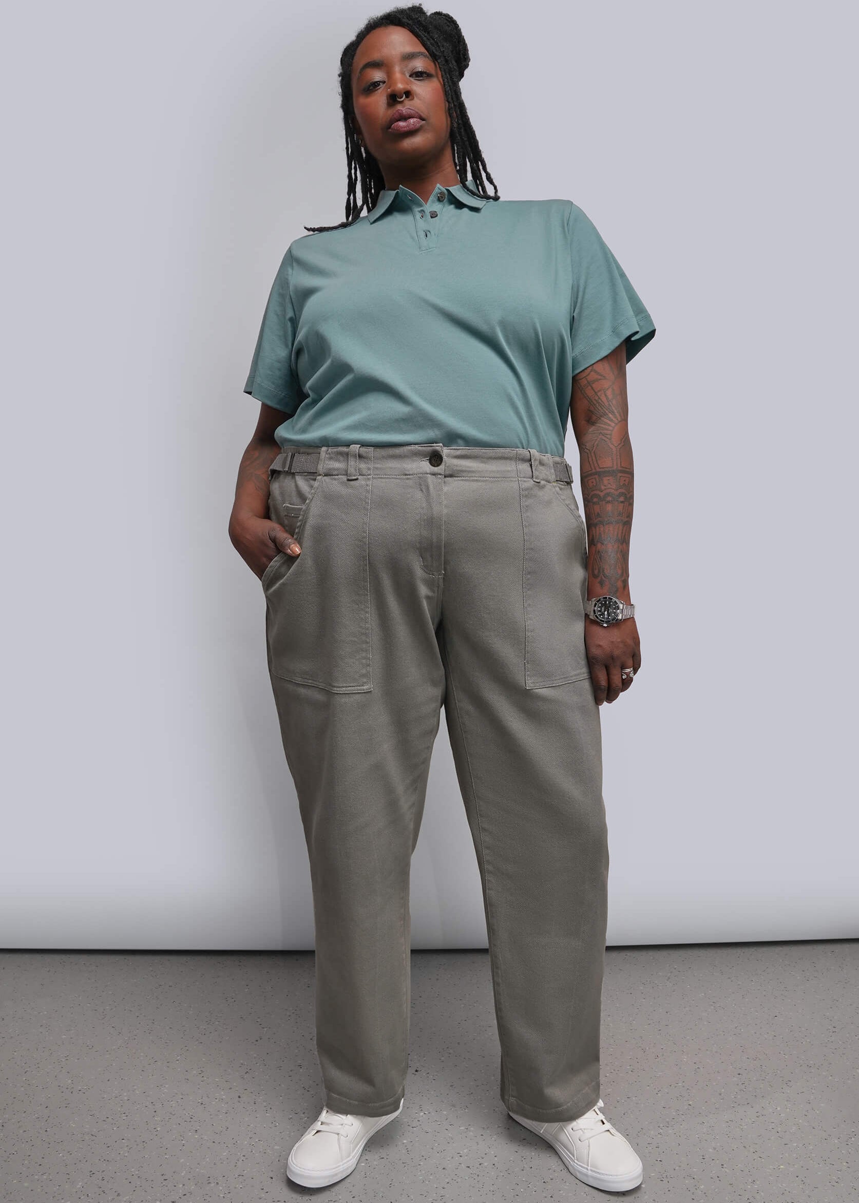 1X=A model stands against a light gray studio wall wearing a teal short-sleeve polo shirt, light gray wide-leg trousers, and white sneakers.
