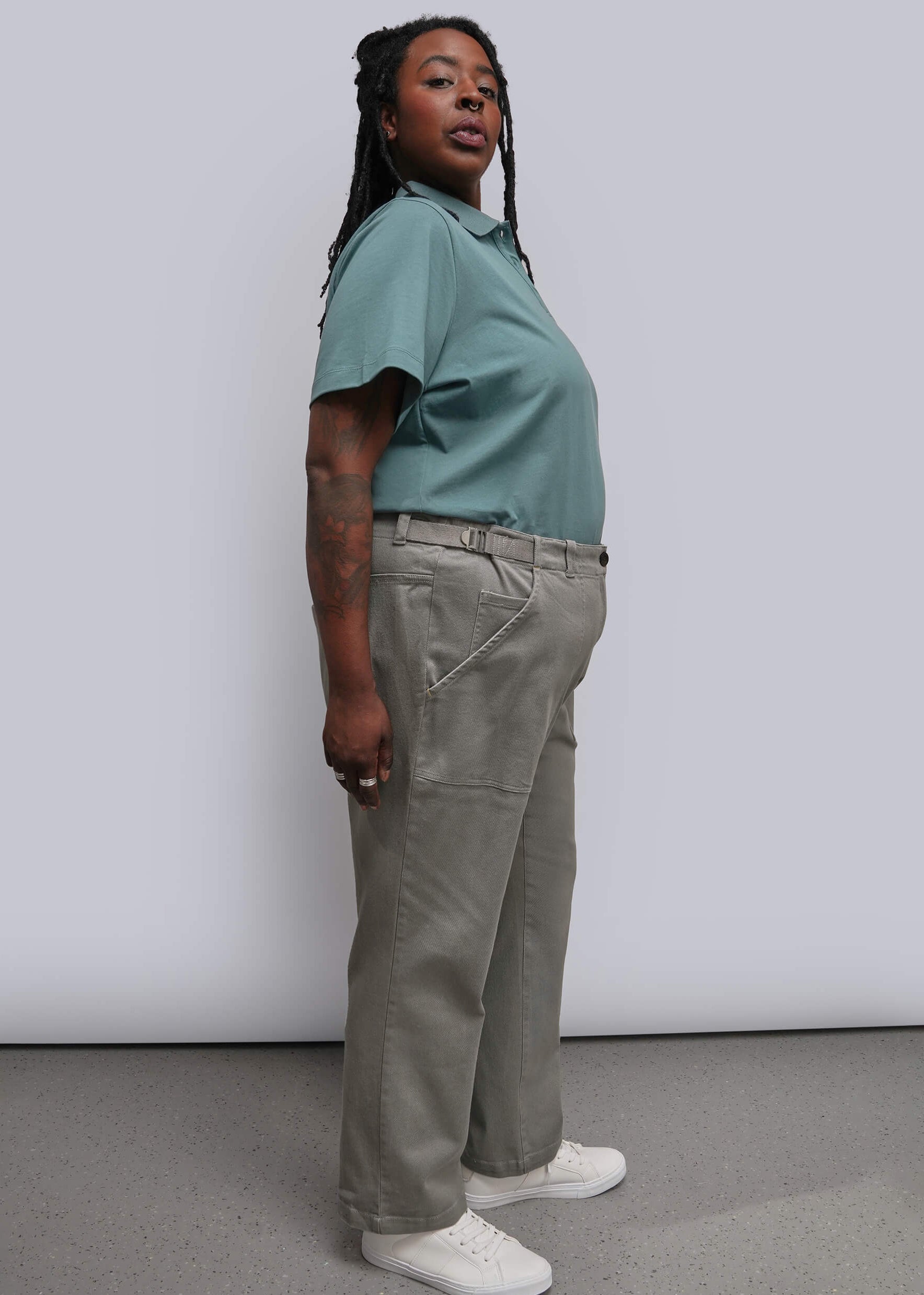 1X=A woman stands in profile wearing a muted sage green short-sleeve polo shirt, light olive-gray wide-leg trousers, and white sneakers against a plain light gray studio wall and floor.