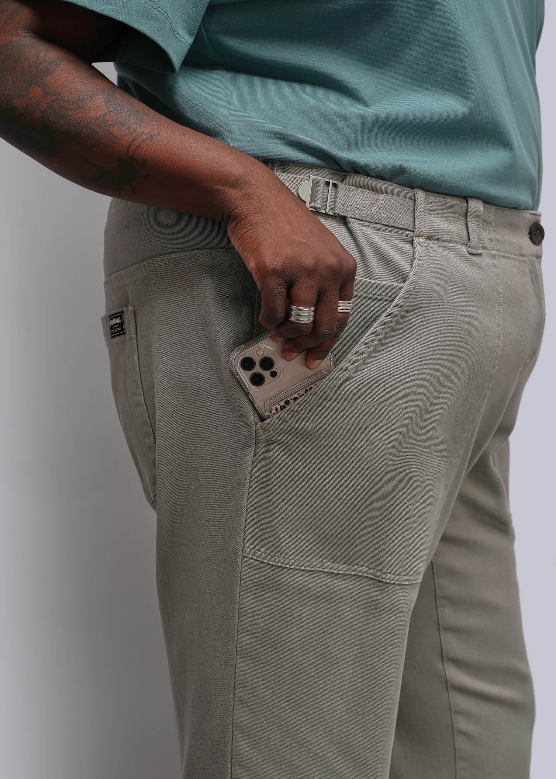 1X=Close-up of a person wearing light gray cargo-style pants with a side pocket holding a phone, paired with a teal short-sleeve top against a plain light gray background.