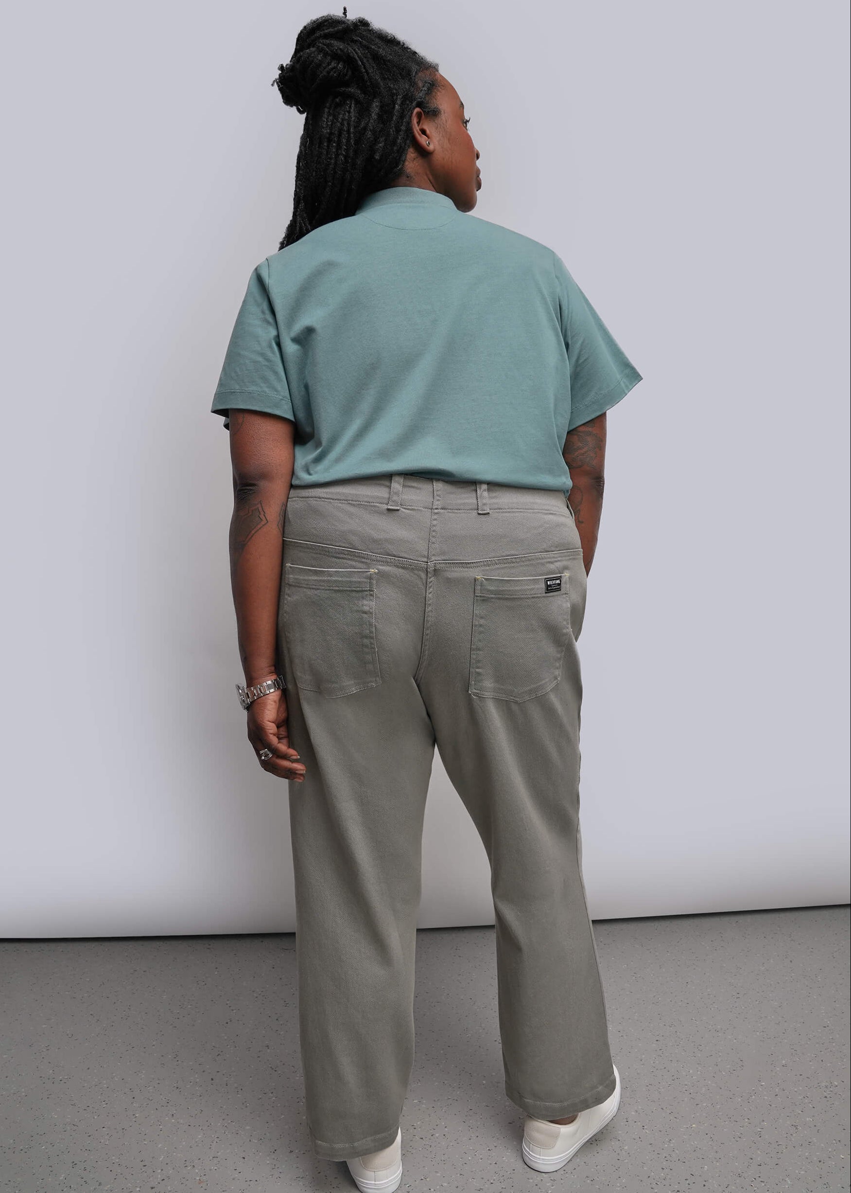 1X=A person shown from the back wearing a teal short-sleeve polo shirt and light olive green wide-leg trousers, paired with white sneakers, against a plain light gray studio background.