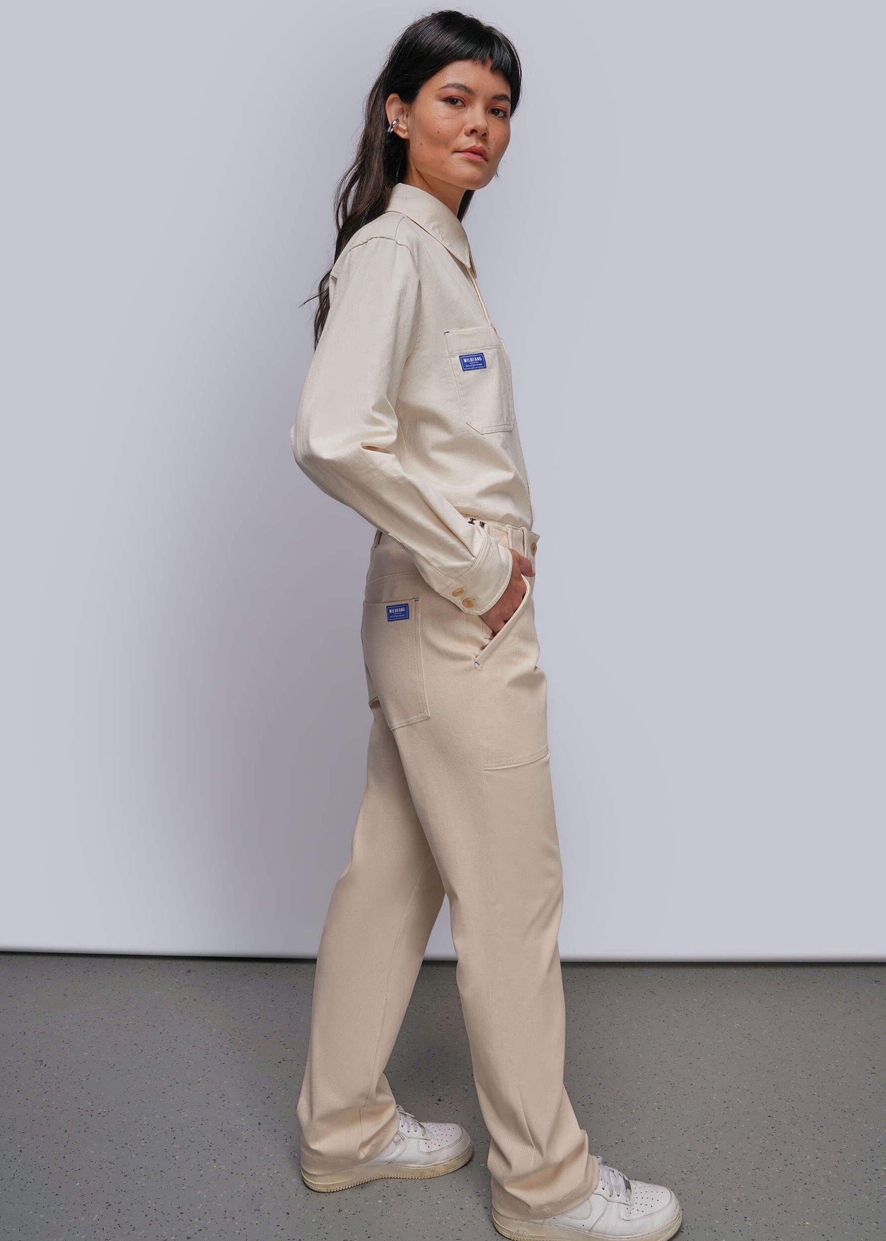 S=A woman modeling a light beige denim matching button up and utility pant paired with white sneakers, set against a plain light gray studio background.