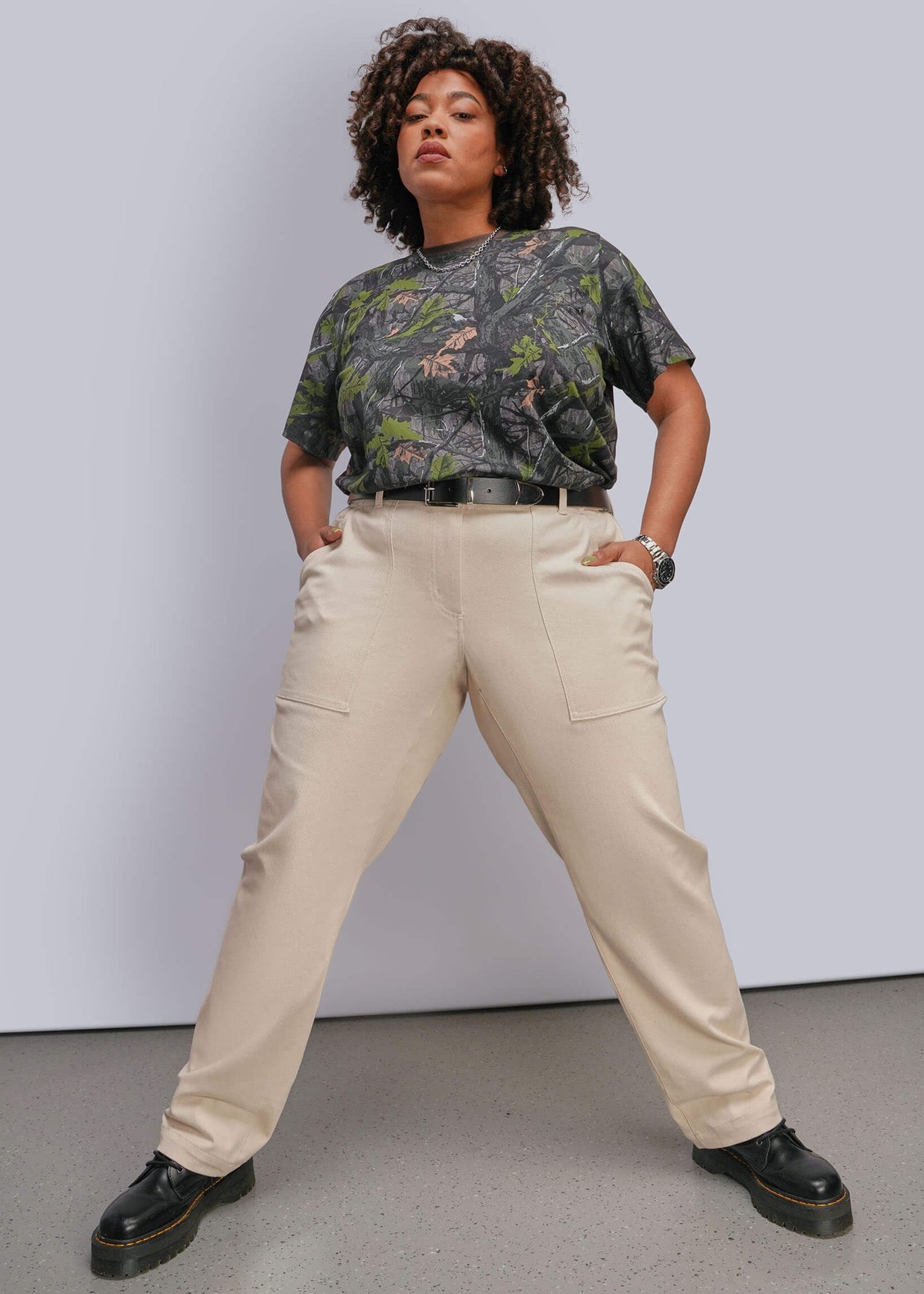 1X=A model wearing a camouflage patterned t-shirt, beige cargo pants, and black leather shoes, posing against a light gray studio wall.