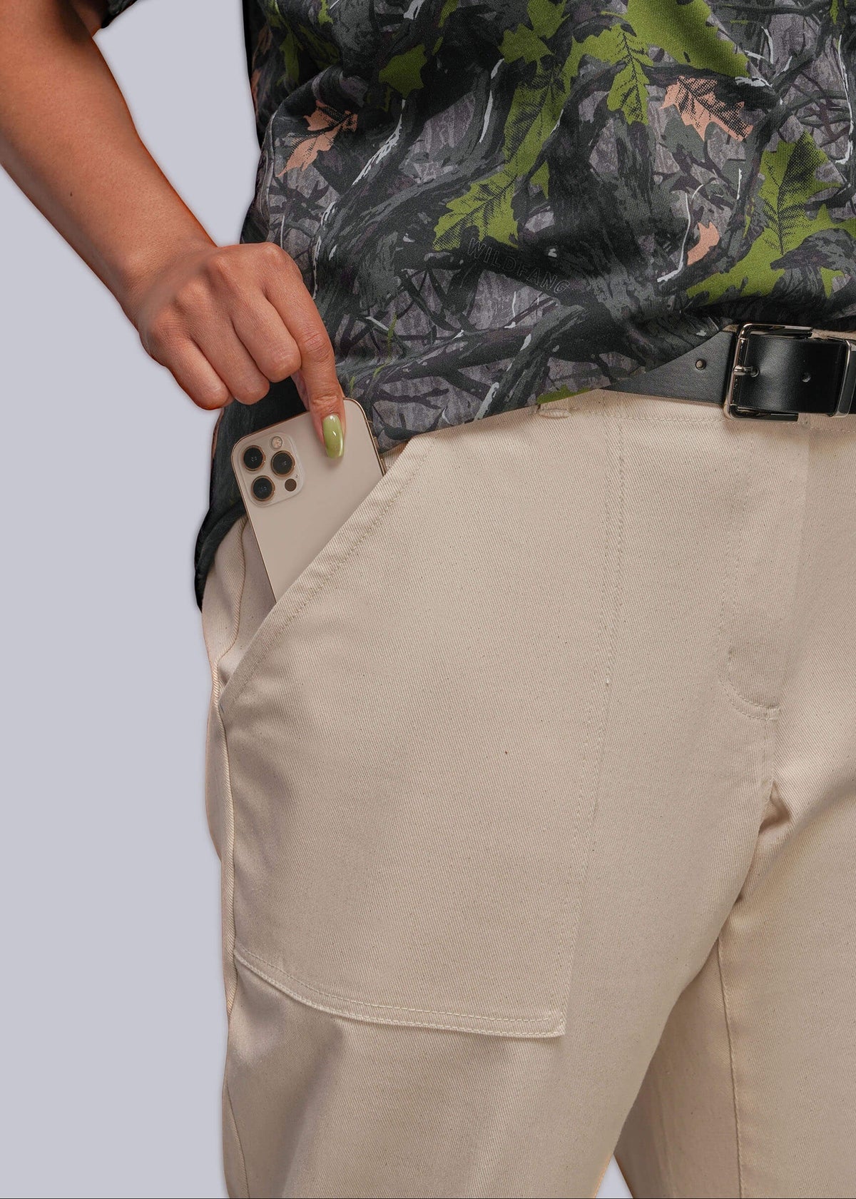 1X=A close-up of a person wearing beige cargo-style pants with a side pocket holding a smartphone, paired with a black belt and a dark camouflage shirt, against a light gray studio background.