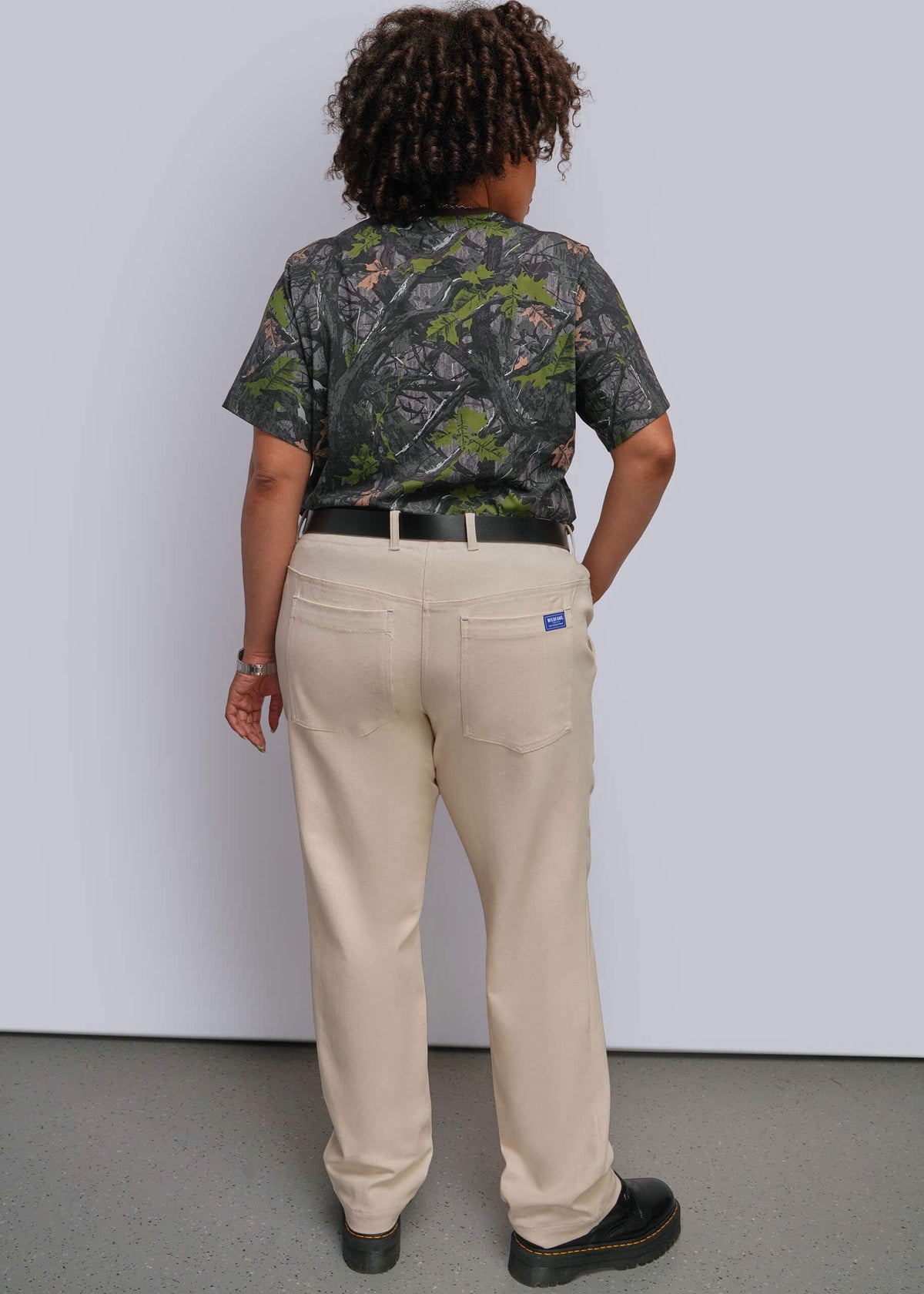 1X=A person shown from the back wearing a short-sleeve camouflage-patterned shirt, beige straight-leg work pants with a black belt, and black shoes, standing against a light gray wall.