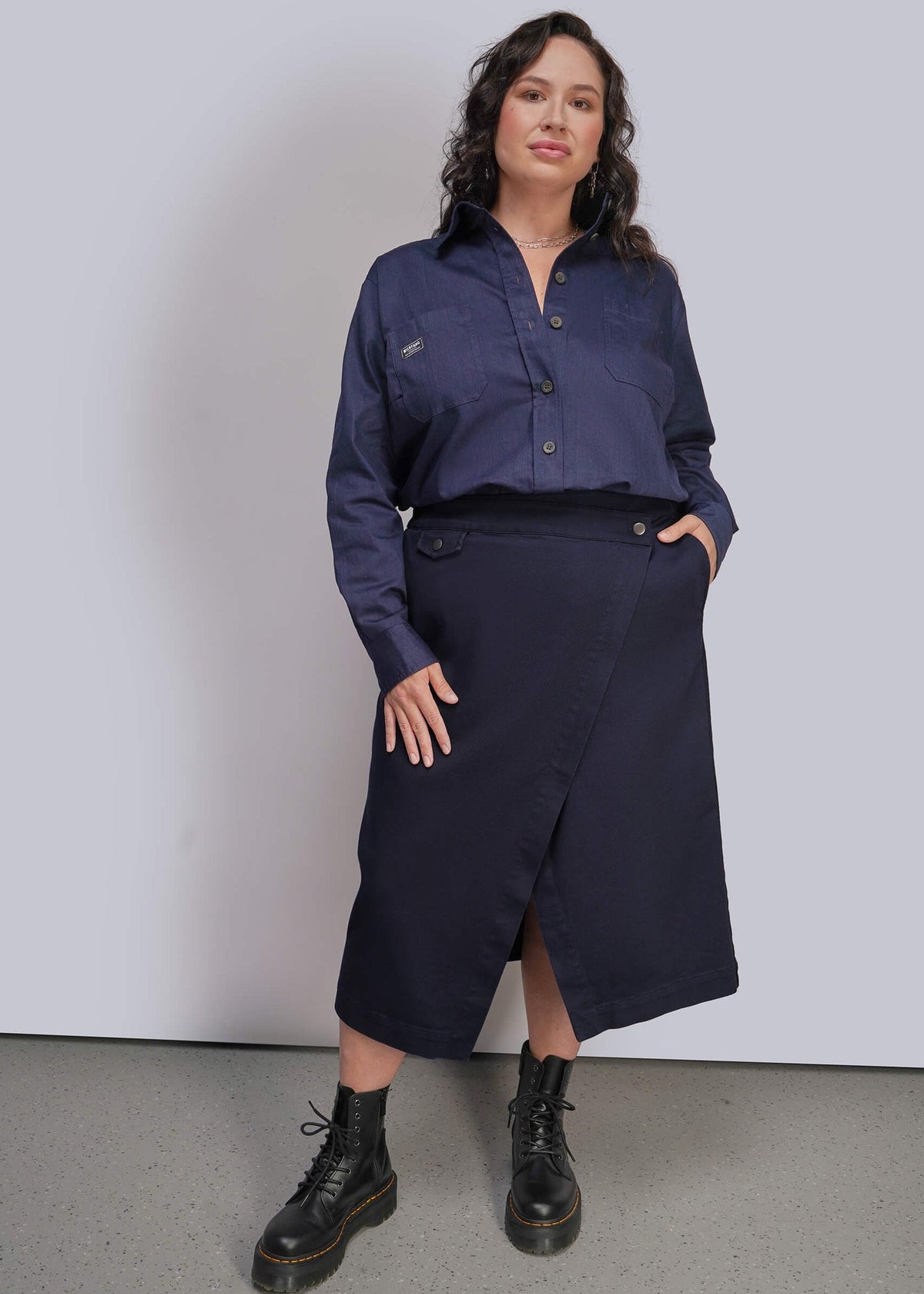 1X=A woman modeling a dark navy blue button up shirt and matching dark navy wrap midi skirt, paired with black combat boots, against a light gray studio background.