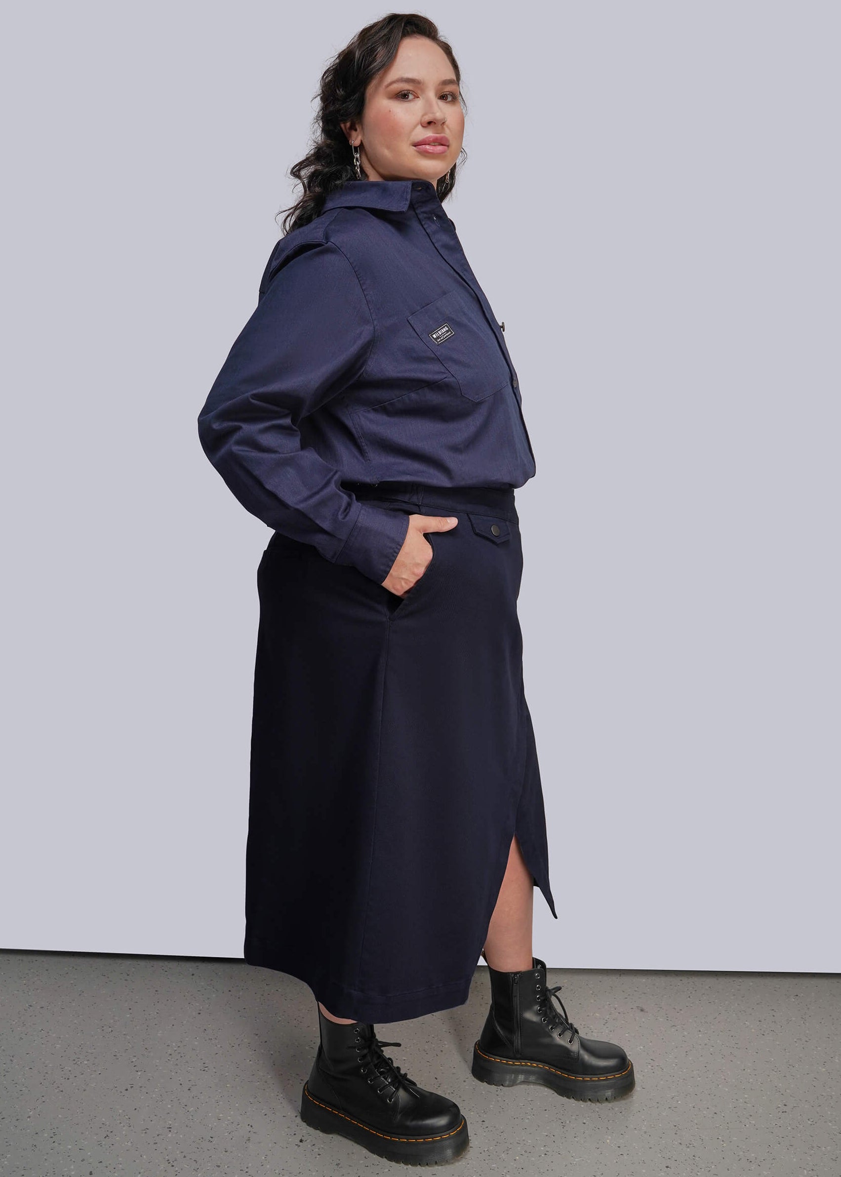 1X=A woman modeling a navy blue button up and matching wrap skirt set, paired with black combat boots, against a light gray studio background.
