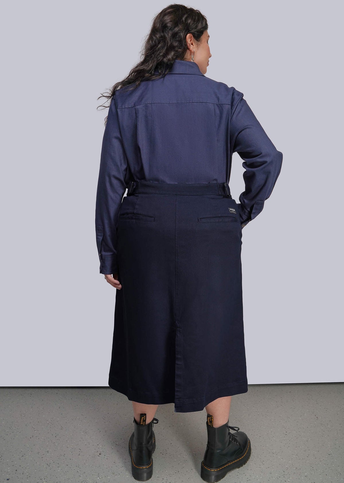 1X=A model is shown from the back wearing a navy blue long-sleeve collared shirt tucked into a matching navy midi skirt, paired with black boots, against a light gray studio background.