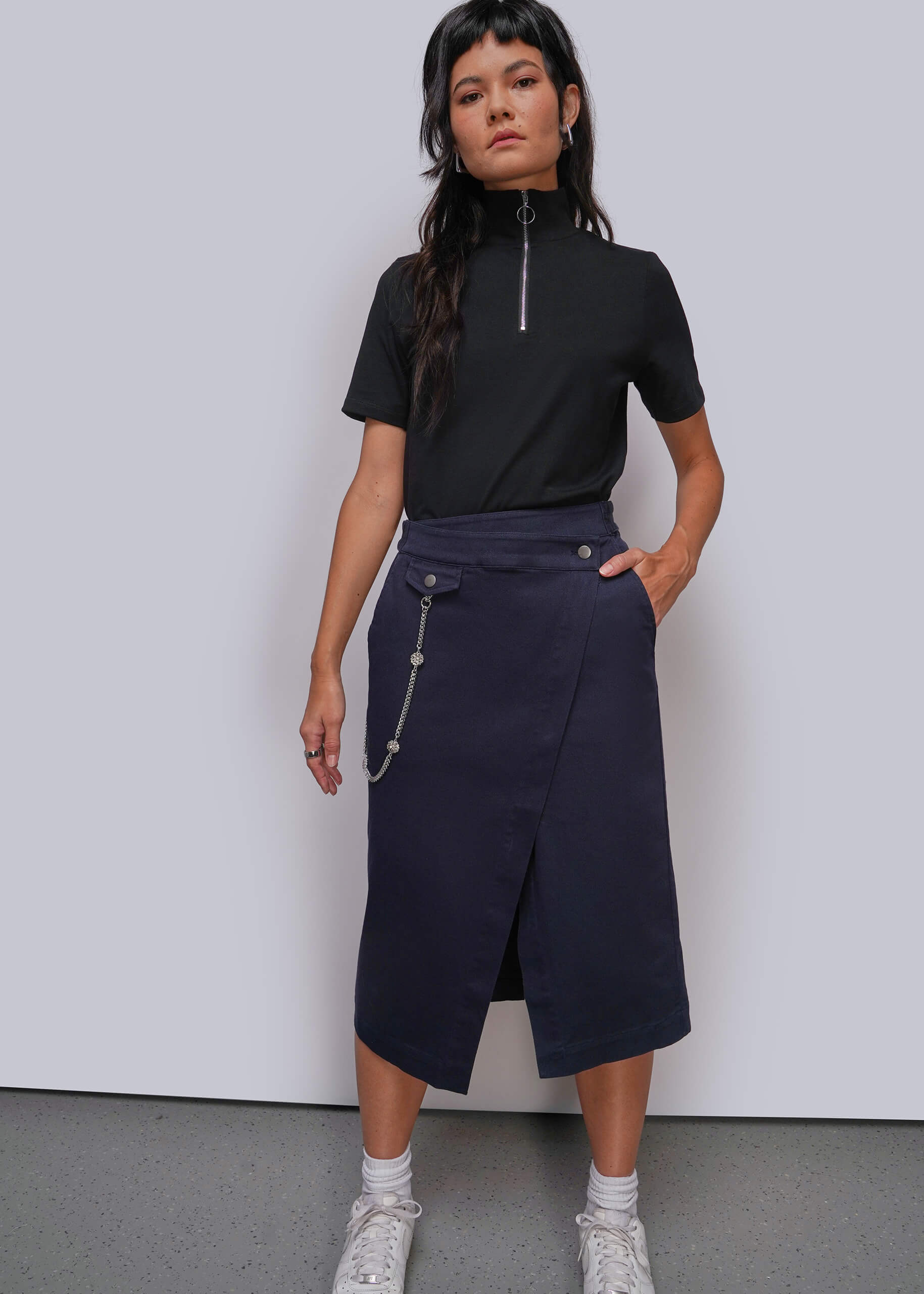 A model stands against a light gray studio wall wearing a black short-sleeve mock-neck top with a silver zipper and a dark navy wrap-style midi skirt with silver chain details. She is also wearing white socks and white sneakers.