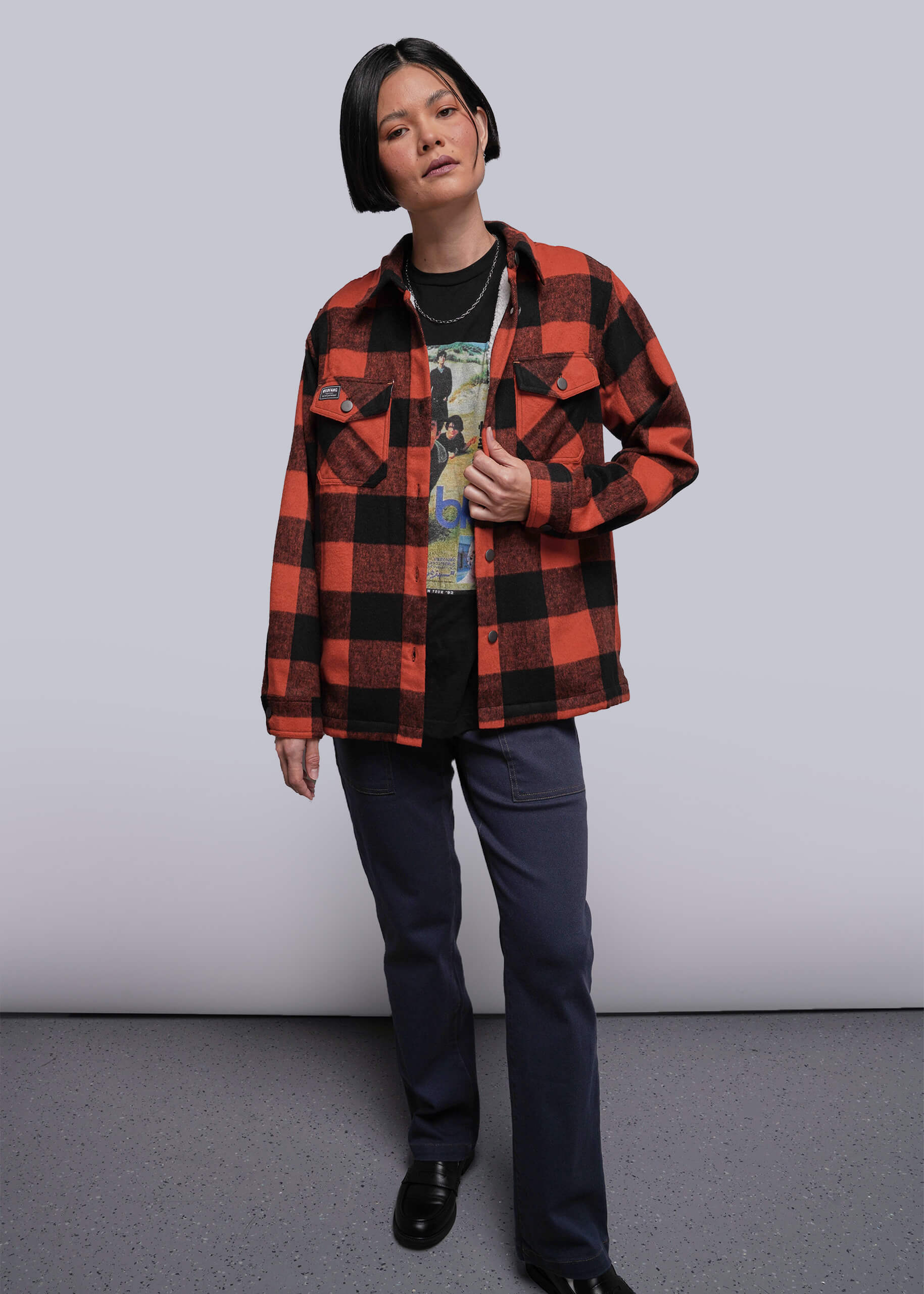 S=Person wearing a red and black plaid shirt over a graphic t-shirt, standing against a plain background.