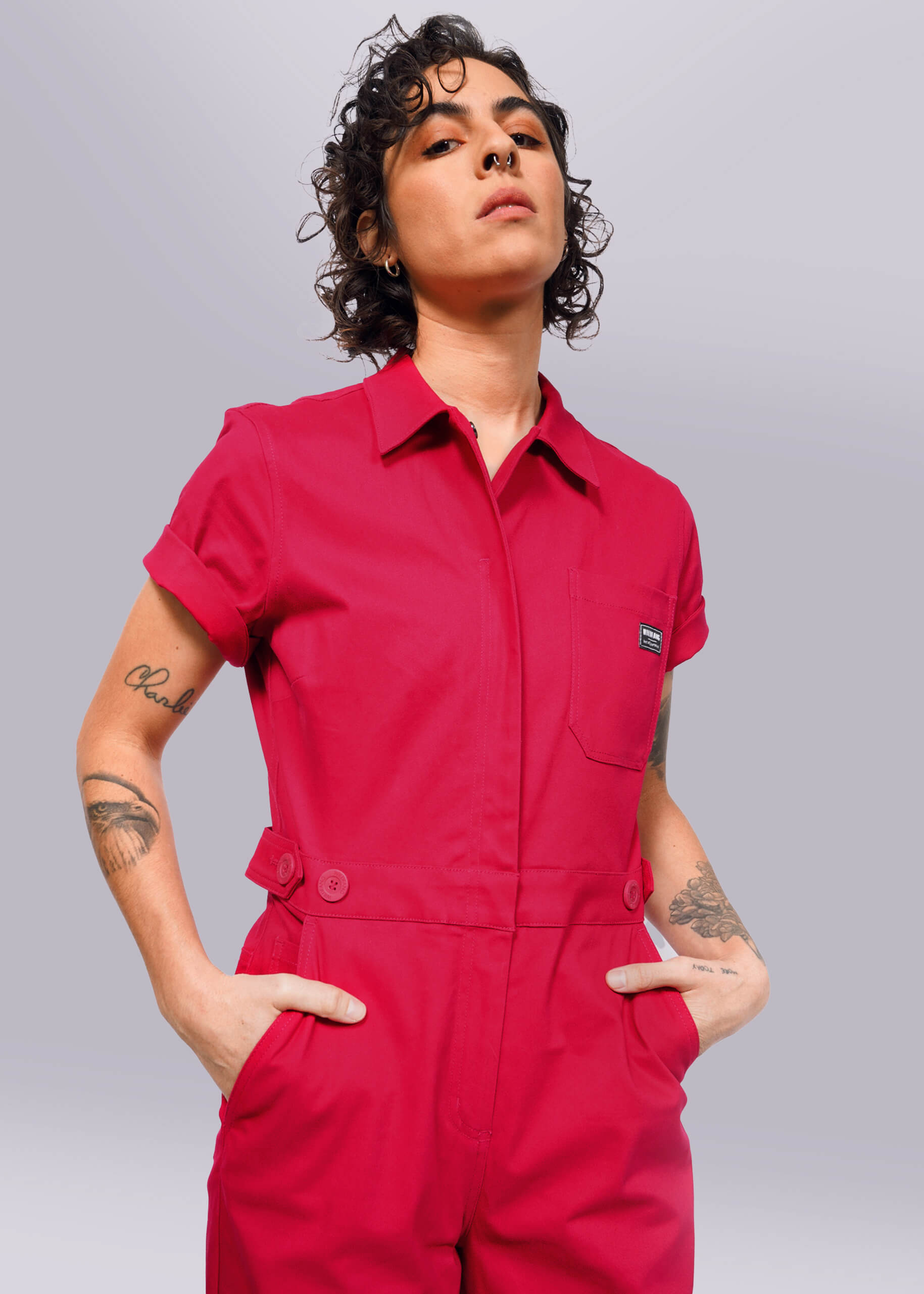 S=Wearing The Essential High Waisted Coverall in bright red, a person stands confidently against a white background. With curly hair and visible tattoos, they have the sleeves rolled up and their hands in deep pockets.