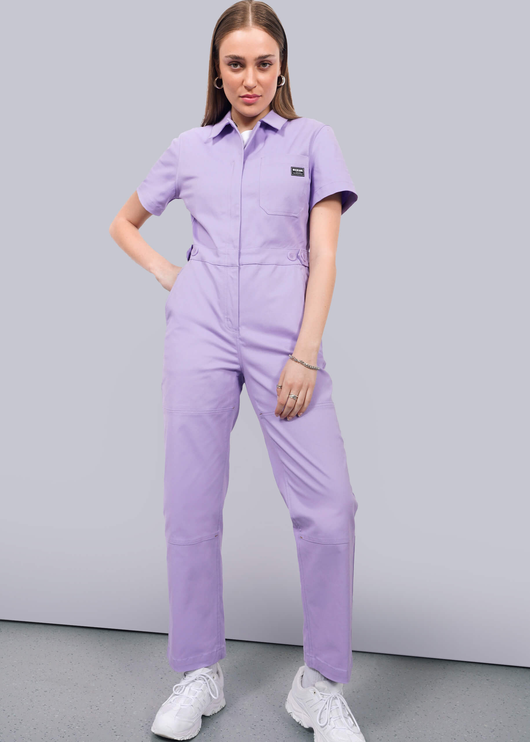 A woman with straight, light brown hair poses against a white background wearing The Essential High Waisted Coverall, a light purple cotton stretch jumpsuit with short sleeves, paired with white sneakers. One hand is on her hip, the other by her side.