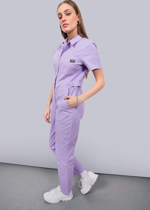 S=A woman stands sideways against a white wall, wearing The Essential High Waisted Coverall in light purple with short sleeves and an adjustable waist. Her hands are in her pockets as she confidently poses, styled with white sneakers.
