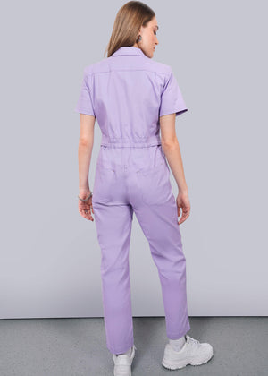 S= A woman with long, light brown hair stands with her back to the camera, wearing The Essential High Waisted Coverall in light purple and white sneakers, facing a white wall on a grey floor.