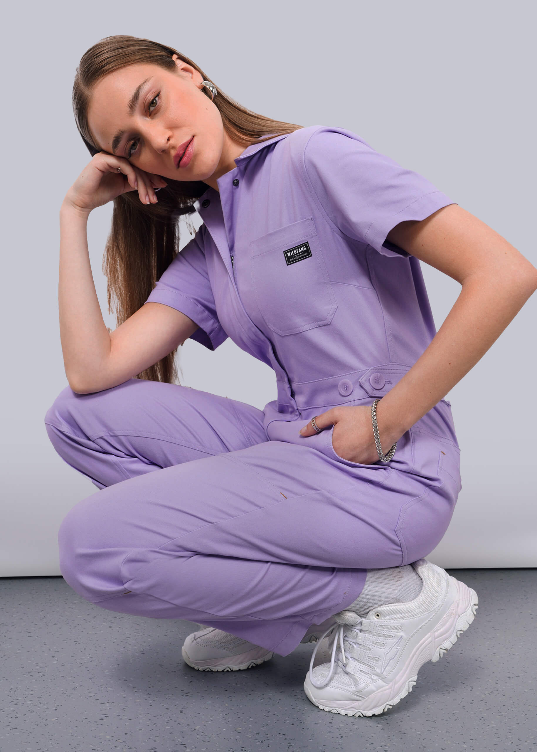A woman wearing The Essential High Waisted Coverall in light purple and white sneakers squats on a gray floor, resting her head on her hand and gazing at the camera with a neutral expression.