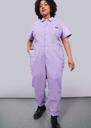 1X= A person with curly hair wears The Essential High Waisted Coverall in light purple with short sleeves, paired with black boots, standing confidently against a plain white background.