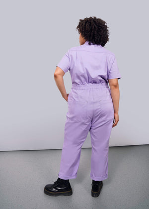 1X= A person with curly hair faces away from the camera, wearing The Essential High Waisted Coverall in lavender with deep pockets and black boots, set against a plain white background.