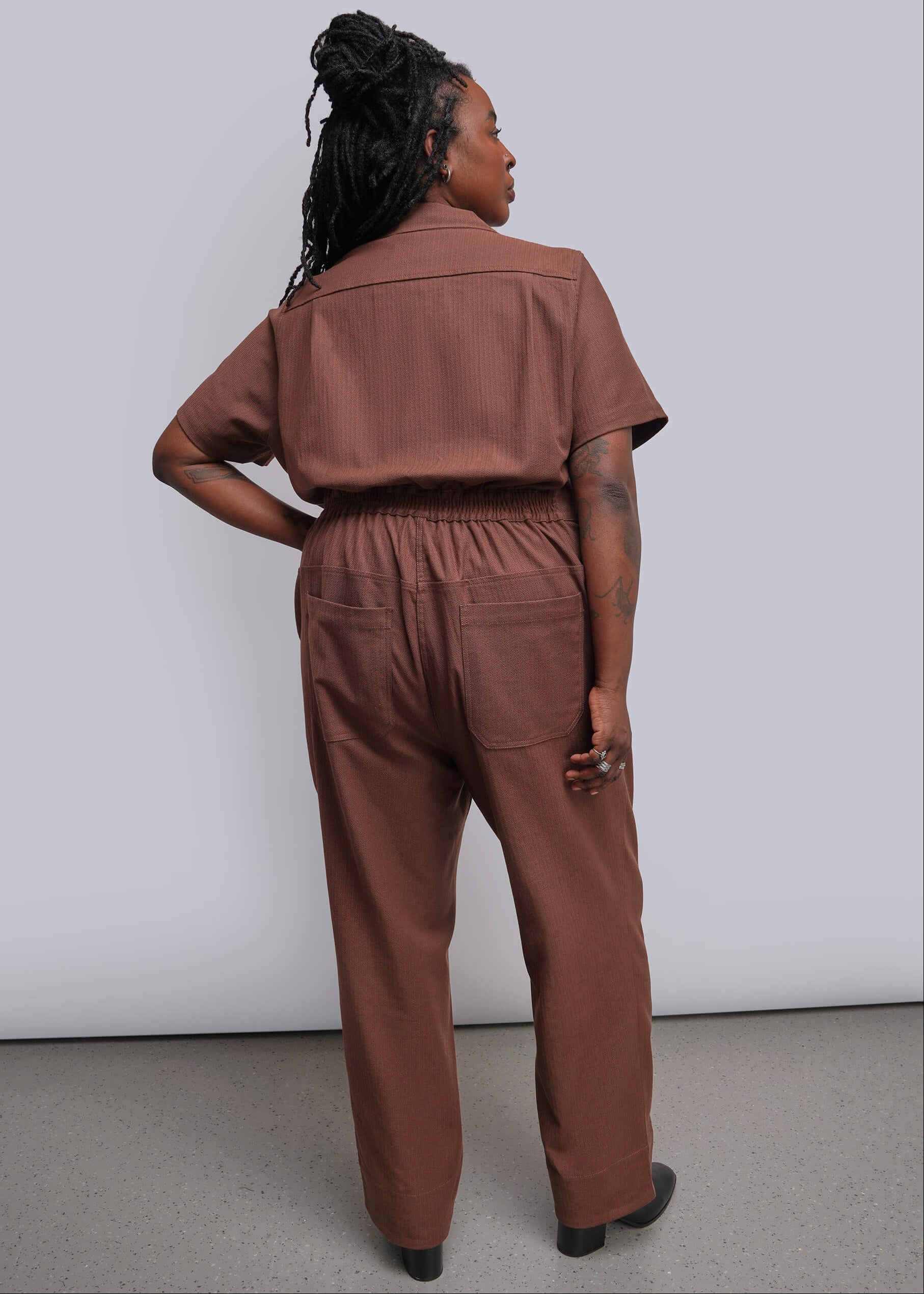 1X=A woman wearing a rust-brown short-sleeve jumpsuit, shown from the back, standing against a plain light gray wall.