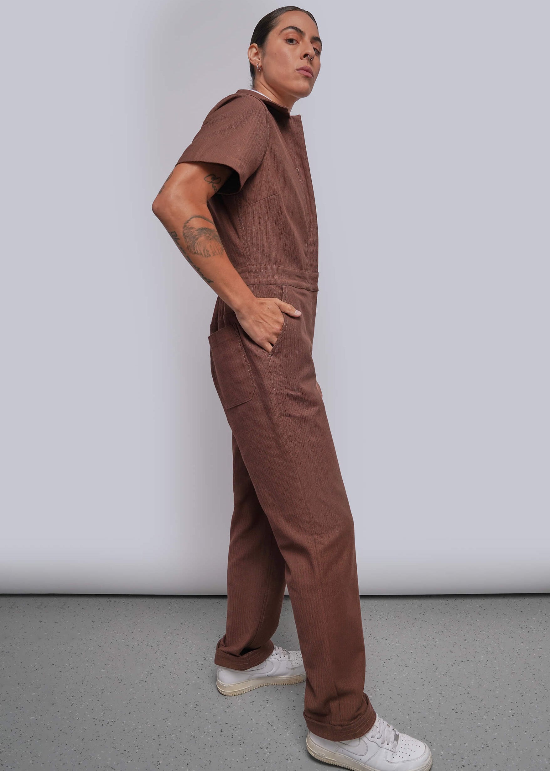 S=A model wearing a brown short-sleeve coverall jumpsuit and white sneakers, standing in profile against a light gray studio background.