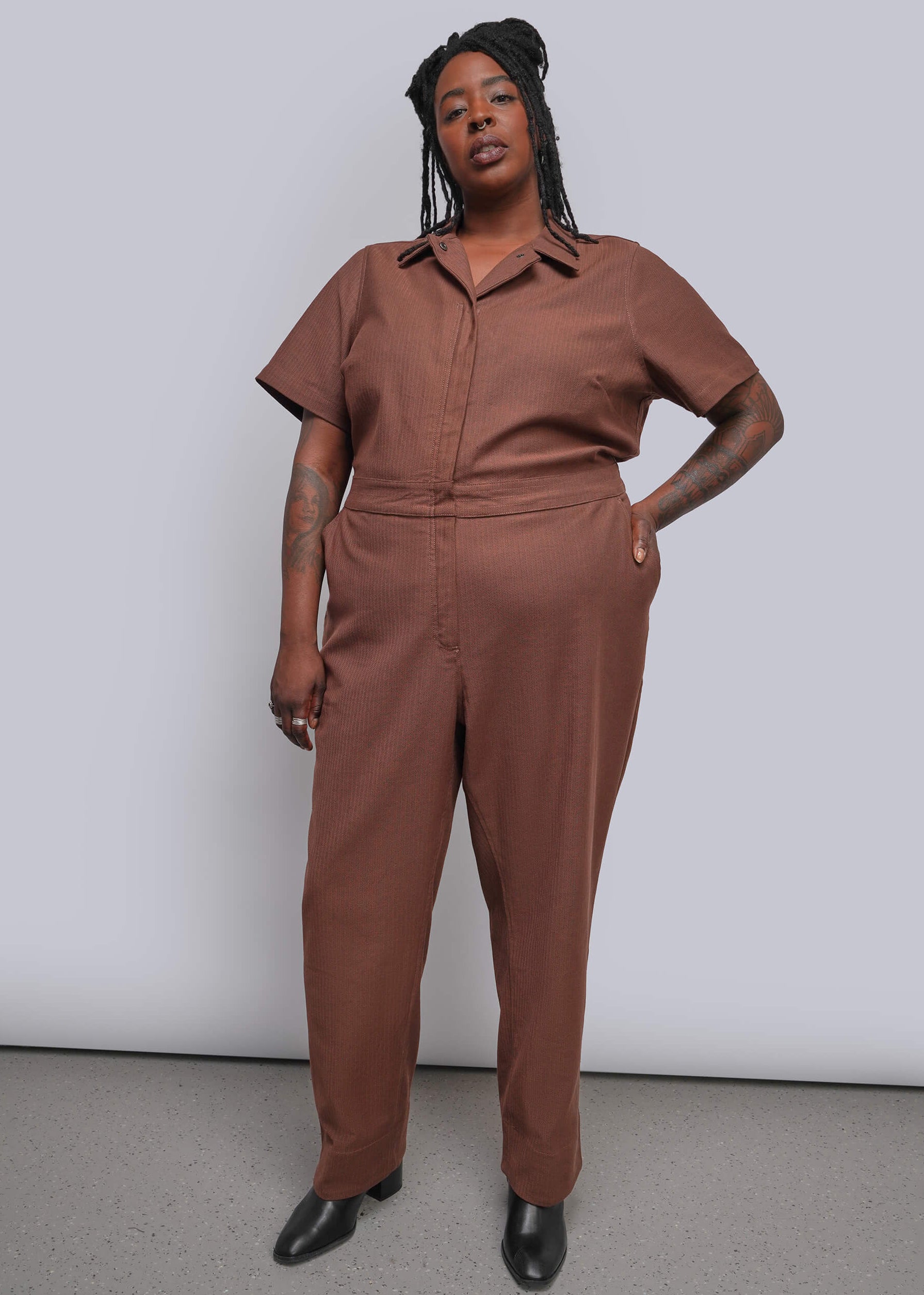 1X=A plus-size model wearing a short-sleeve, button-front jumpsuit in a warm brownish-terracotta color, paired with black pointed shoes, standing against a light gray studio background.