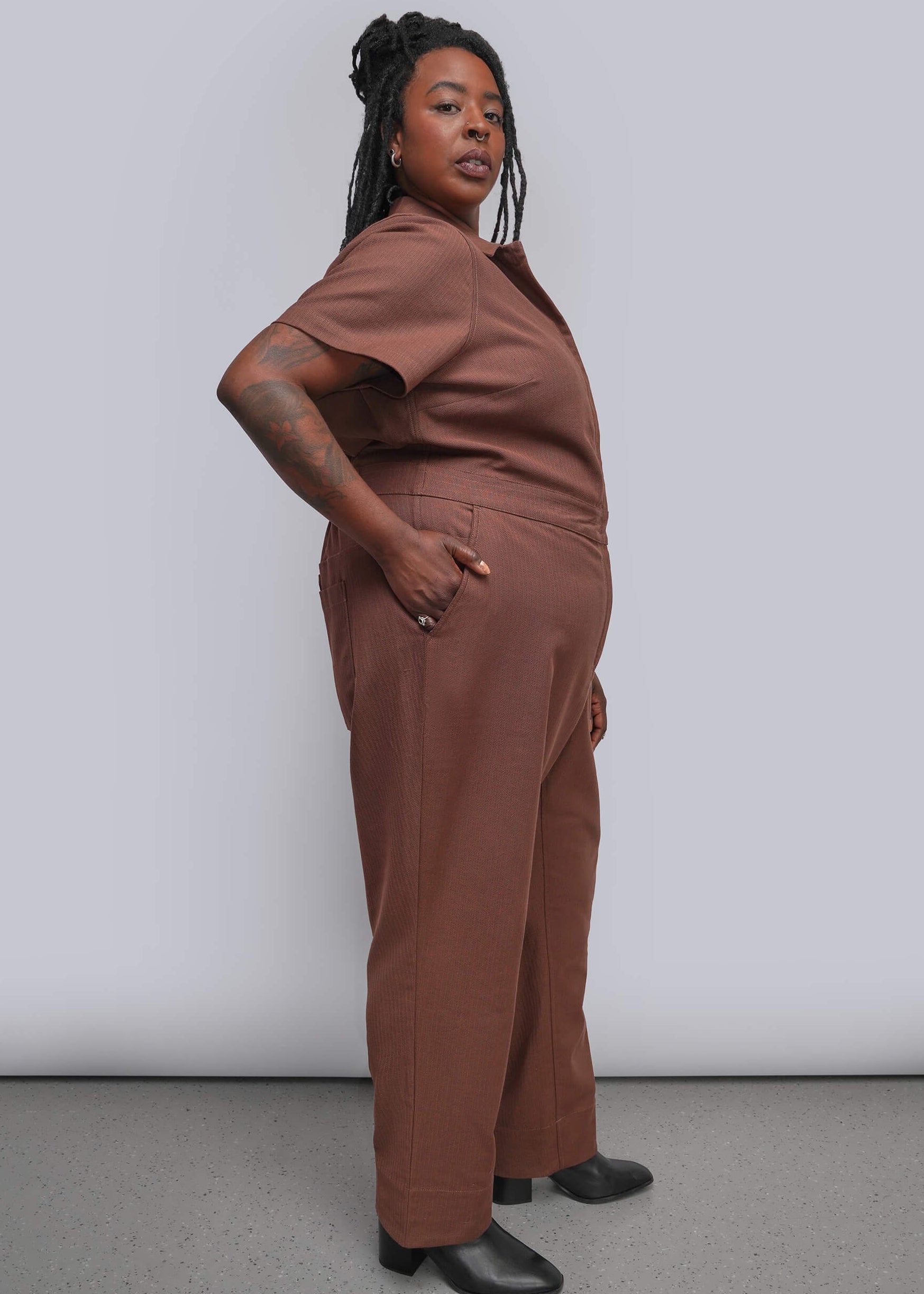 1X=A woman modeling a matching two-piece outfit consisting of a short-sleeve collared top and wide-leg trousers in a dusty terracotta brown color, paired with black shoes, against a plain light gray studio background.