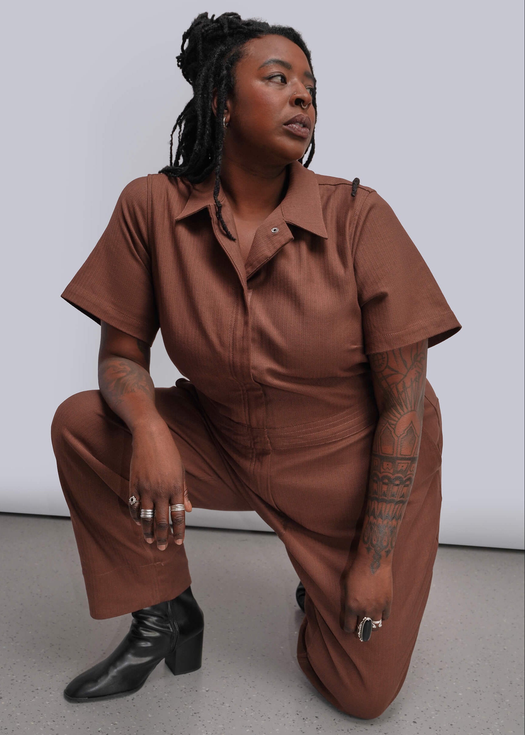 1X=A model wearing a rust-brown short-sleeve jumpsuit and black leather boots, crouching in a studio setting with a light gray background.