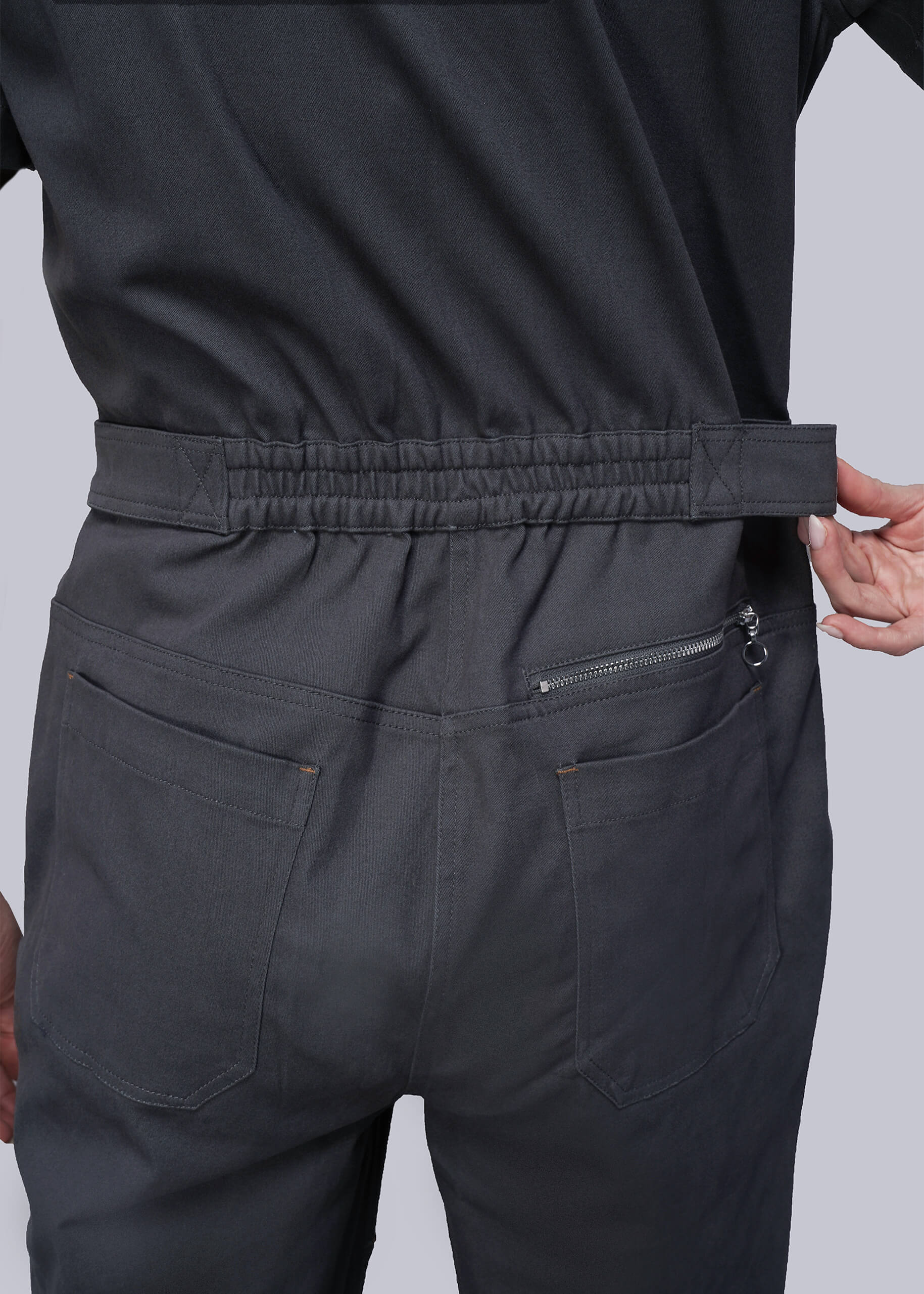 S=Close up of person wearing dark grey jumpsuit with zipper pocket on a light gray background