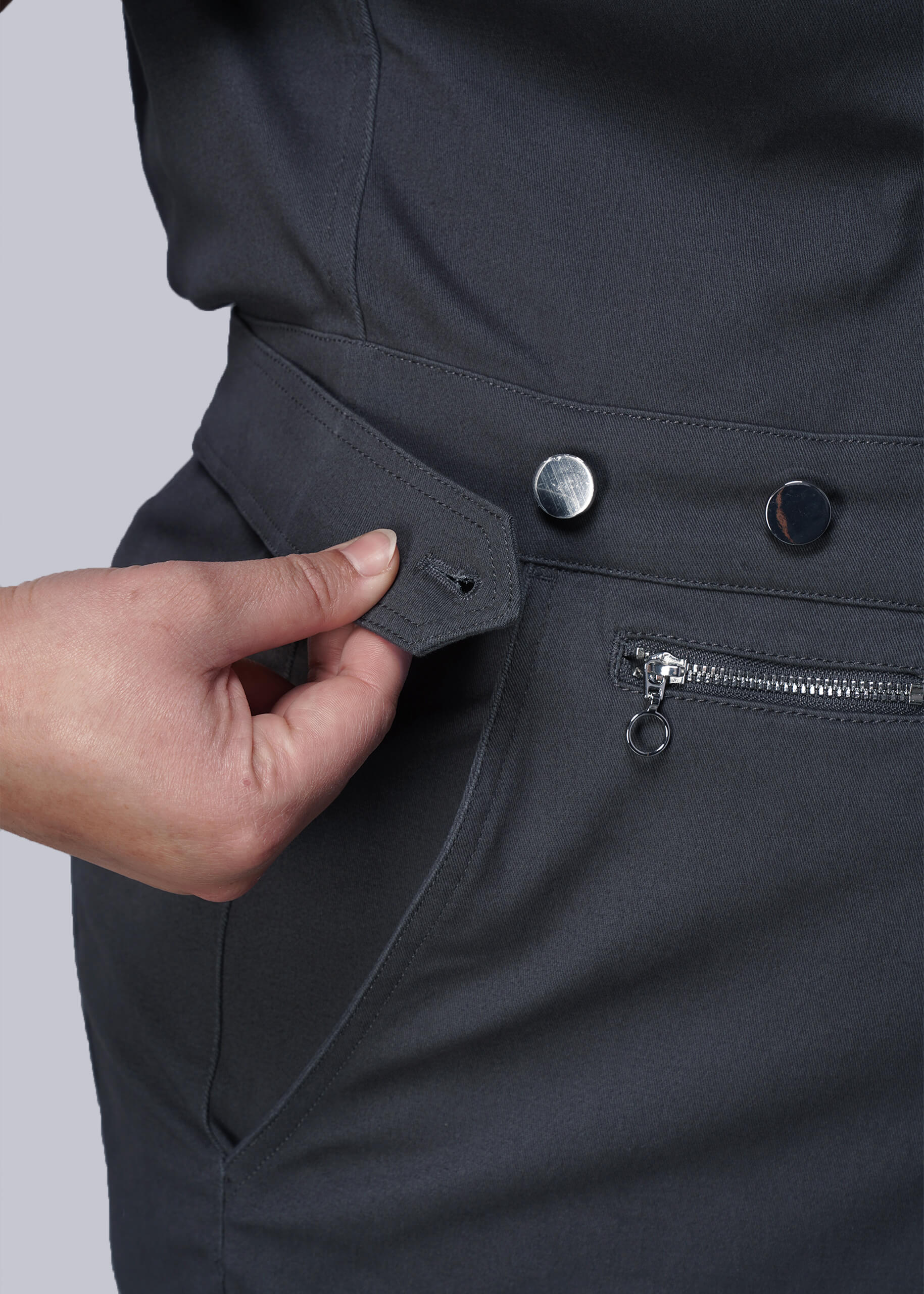 1X=Close-up of a person adjusting an adjustable waist button on a dark grey jumpsuit against a light gray background