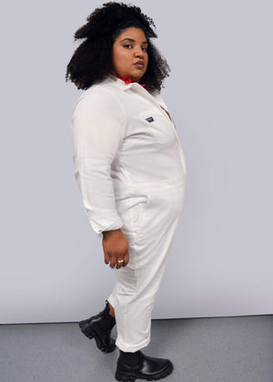 1X=A person in The Essential Long Sleeve Herringbone Coverall, styled with black boots and a red scarf, stands against a gray background. Their curly hair frames their face as they look sideways, adding brightness to the minimalist look.