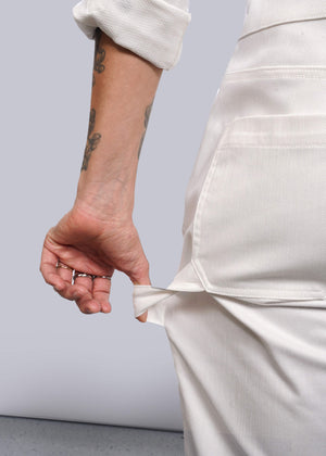 S=A tattooed person wearing The Essential Long Sleeve Herringbone Coverall in white, with the sleeve rolled up, shows the inside of an empty pocket. Their forearm and hand with rings appear against a plain white background.