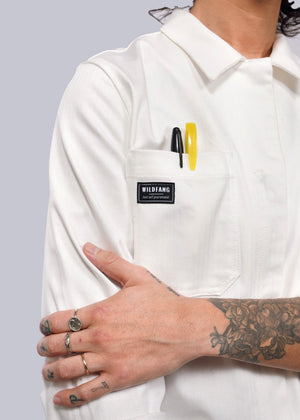 S= In a white Wildfang Essential Long Sleeve Herringbone Coverall, featuring herringbone cotton and 10 pockets, a person stands confidently. Pens peek from one pocket. Tattoos cover an arm, while gleaming rings adorn fingers as they cross arms in front of their body.