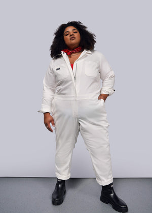 1X= A curly-haired individual stands confidently against a plain background wearing The Essential Long Sleeve Herringbone Coverall, featuring a full-length zipper and multiple pockets for practicality, paired with black boots and accessorized with a red bandana around their neck.