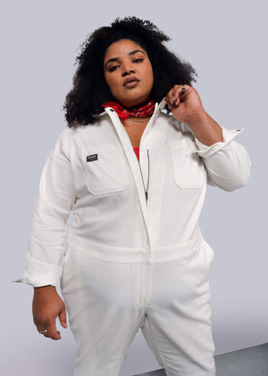 1X=A person with curly hair wears The Essential Long Sleeve Herringbone Coverall, crafted from white cotton, and a red bandana around their neck. They adjust the full-length zipper of the coverall against a plain, light-colored background.