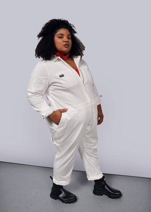 1X= A person with curly hair stands confidently in The Essential Long Sleeve Herringbone Coverall, a white cotton jumpsuit with a full-length zipper. They wear black boots and a red bandana around their neck against a plain grey background.