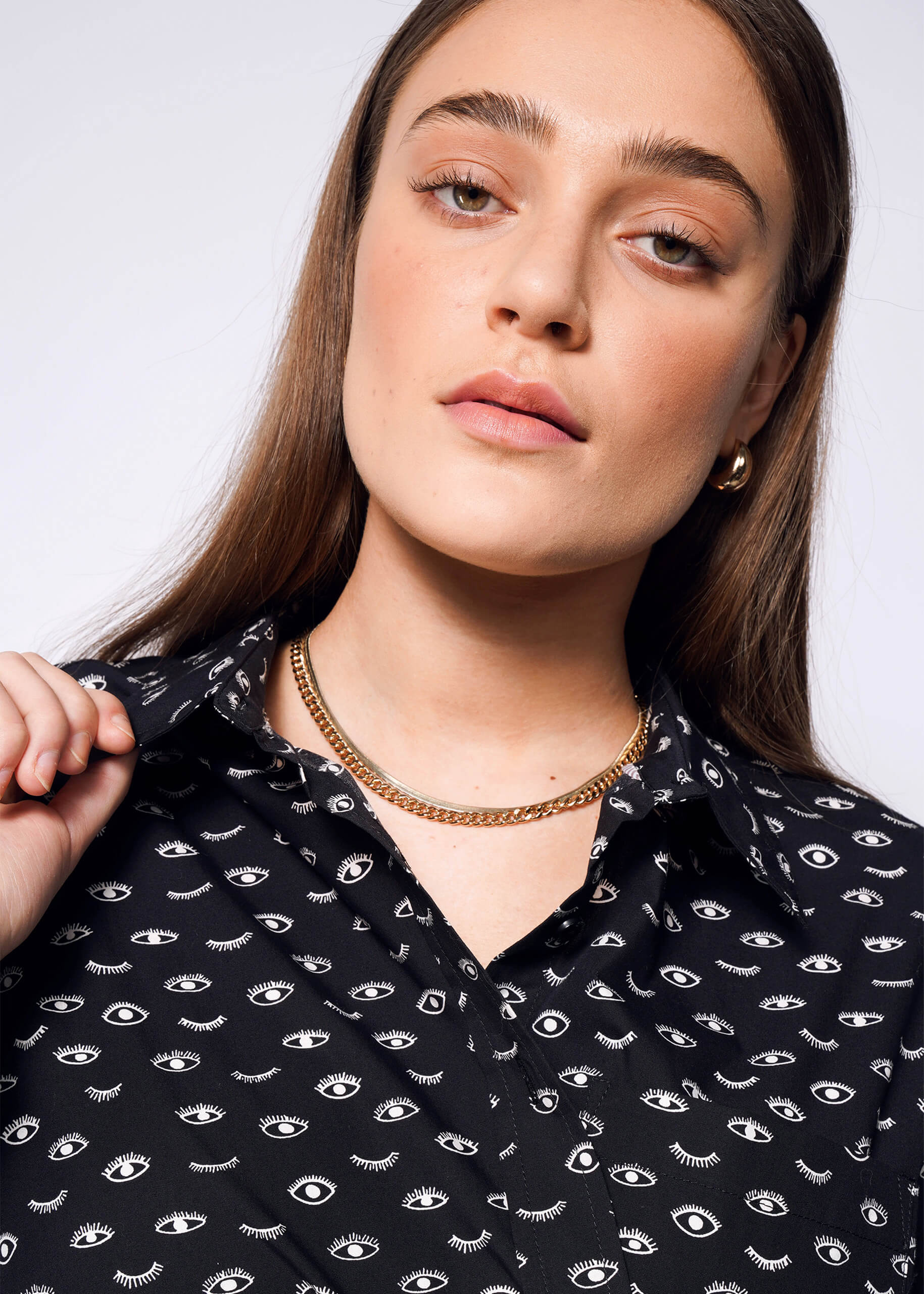 A person with long hair wears The Essential Long Sleeve Oversized Wrap Button Up, featuring white eye patterns on black. Holding the collar, they display a neutral expression, complemented by gold hoop earrings and a layered gold necklace against a plain white background.