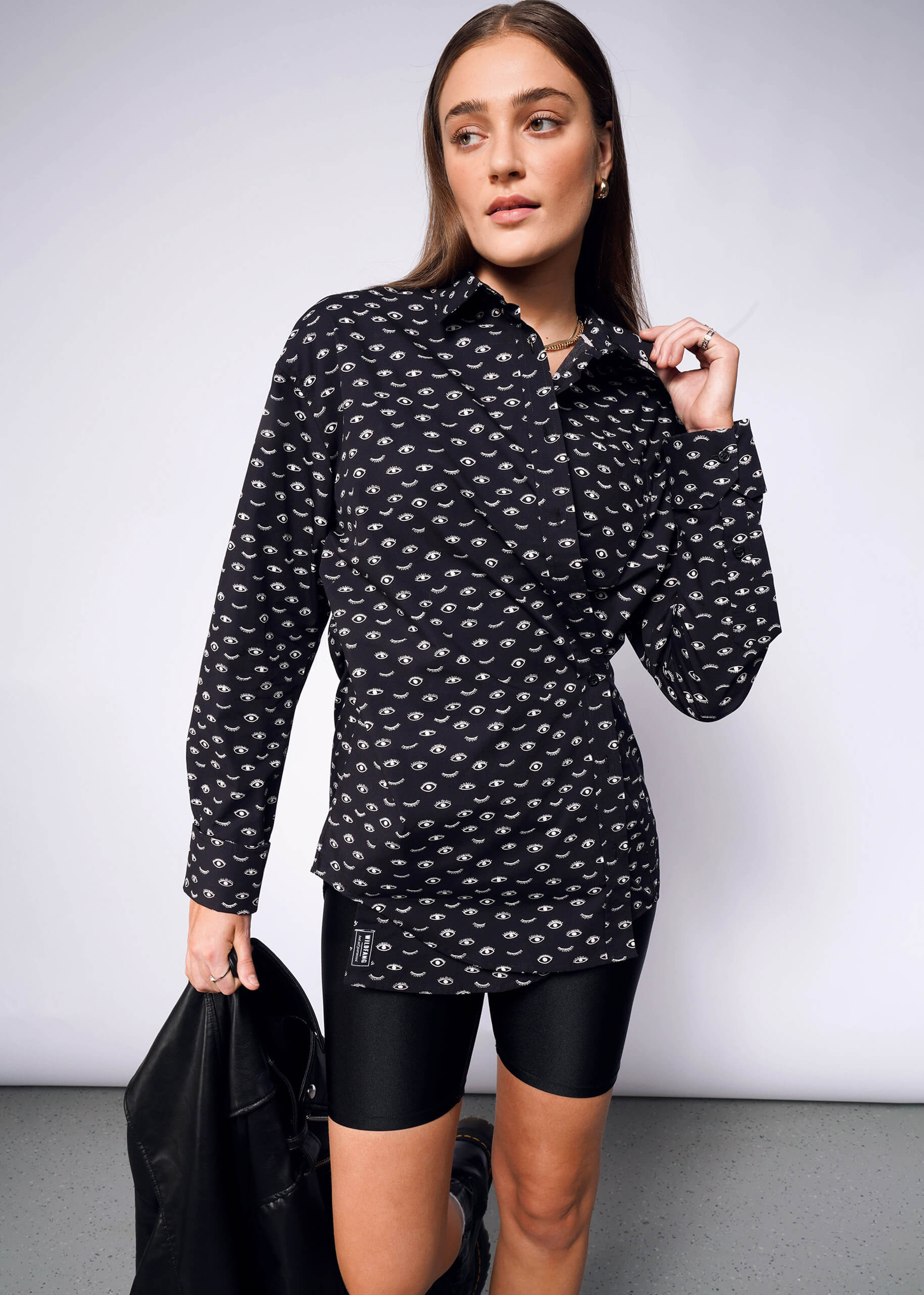 A woman poses confidently, looking left against a plain background, wearing The Essential Long Sleeve Oversized Wrap Button-Up in black with a white pattern. She pairs it with black biker shorts and holds a convertible wrap-style jacket.