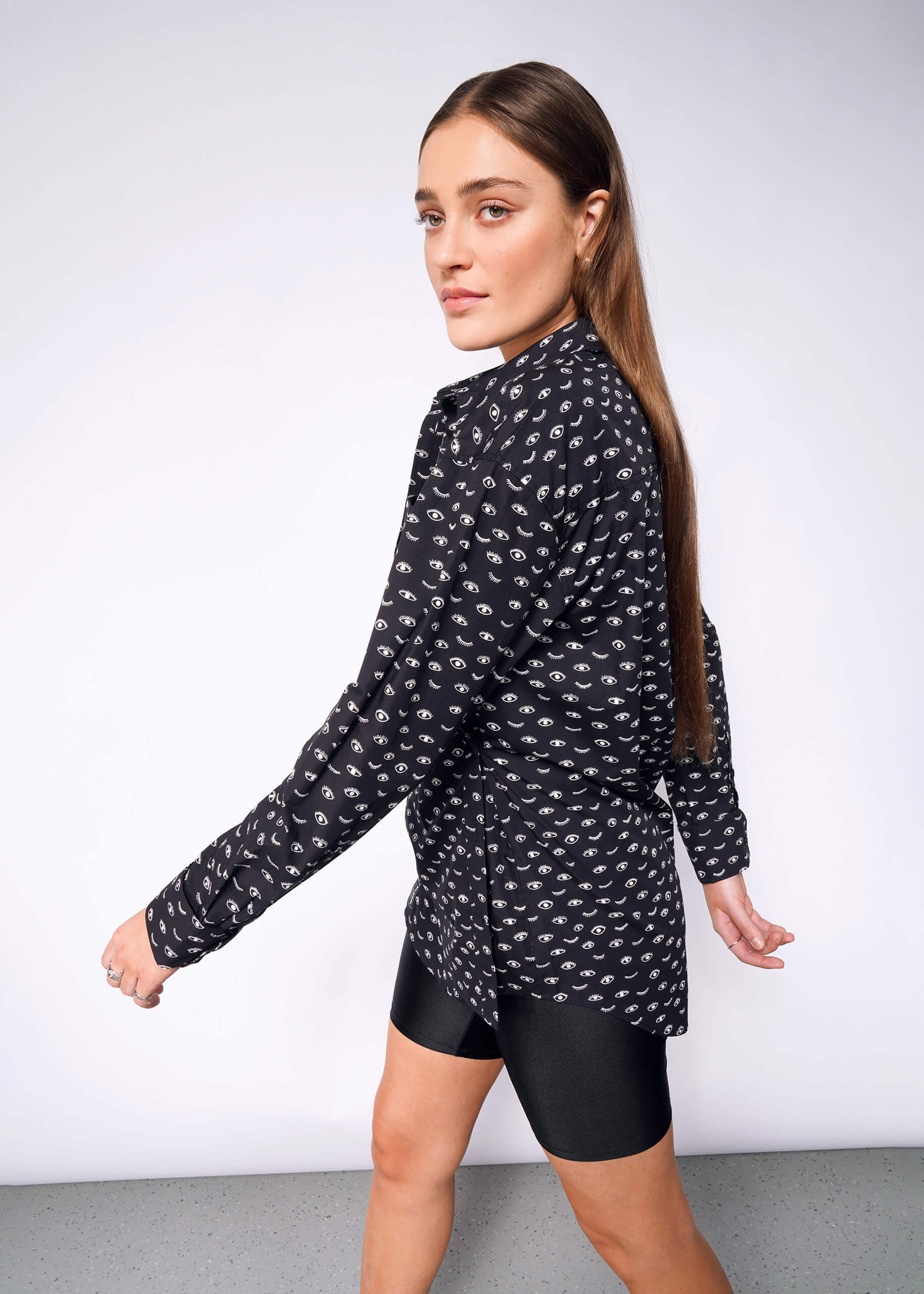 A woman with long brown hair wears The Essential Long Sleeve Oversized Wrap Button Up and black shorts. She walks left, glancing over her shoulder at the camera against a plain gray background.