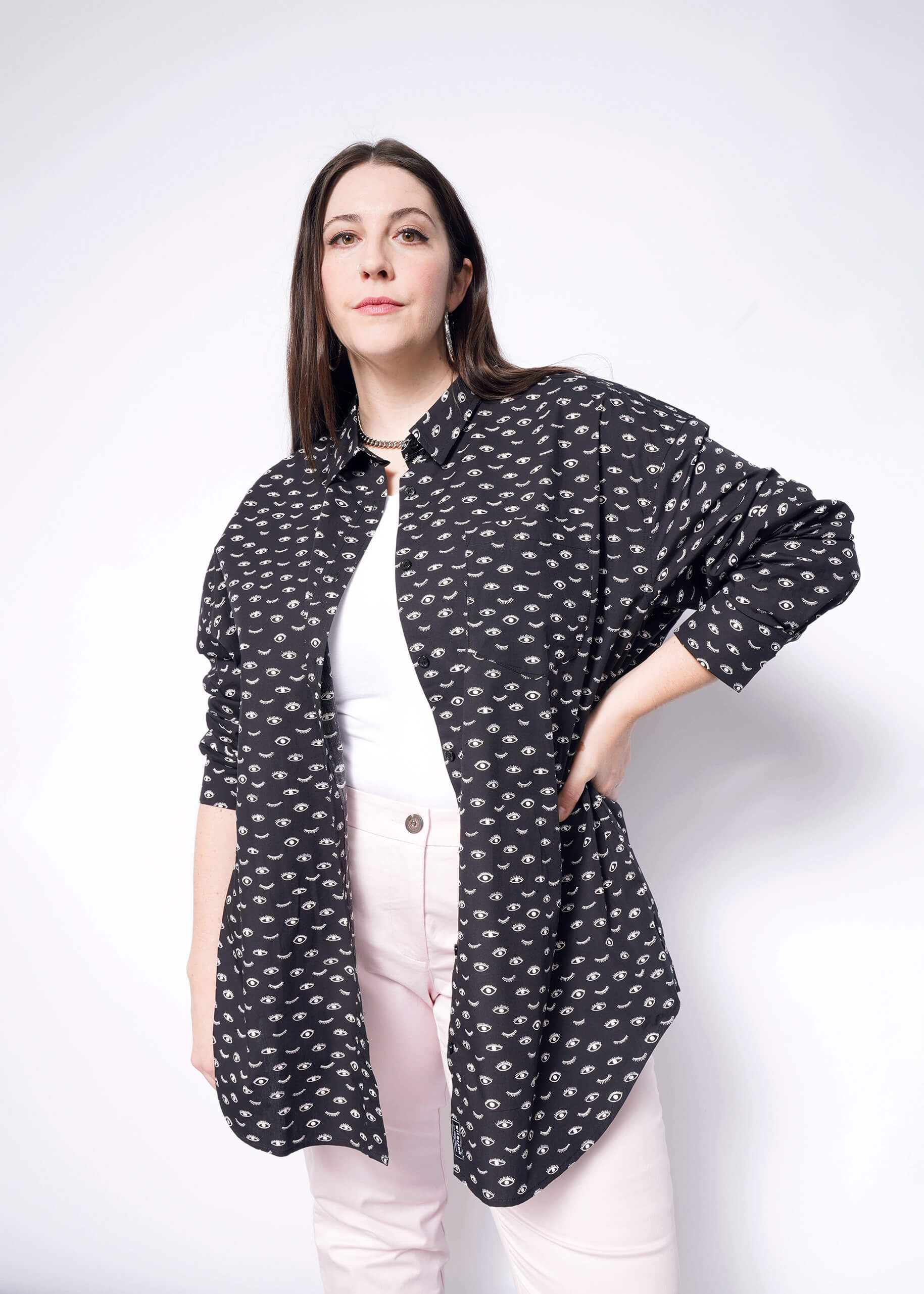 A woman with long brown hair stands against a white background wearing The Essential Long Sleeve Oversized Wrap Button Up in black over a white top, paired with light pink pants. She poses confidently with one hand on her hip and gazes at the camera.