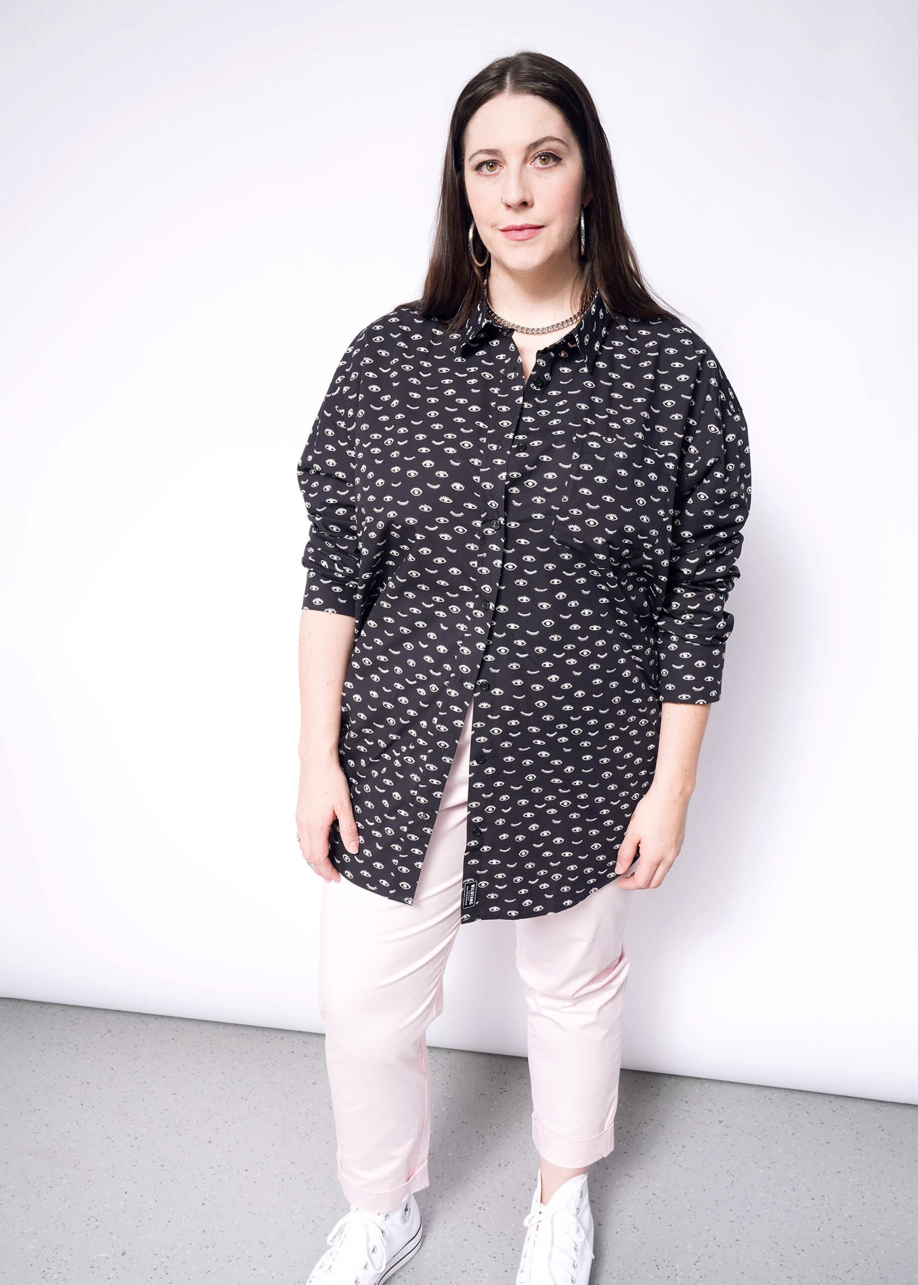 A woman with long, dark hair stands against a white background. Shes dressed in The Essential Long Sleeve Oversized Wrap Button Up shirt featuring a pattern, light pink pants, and white sneakers.