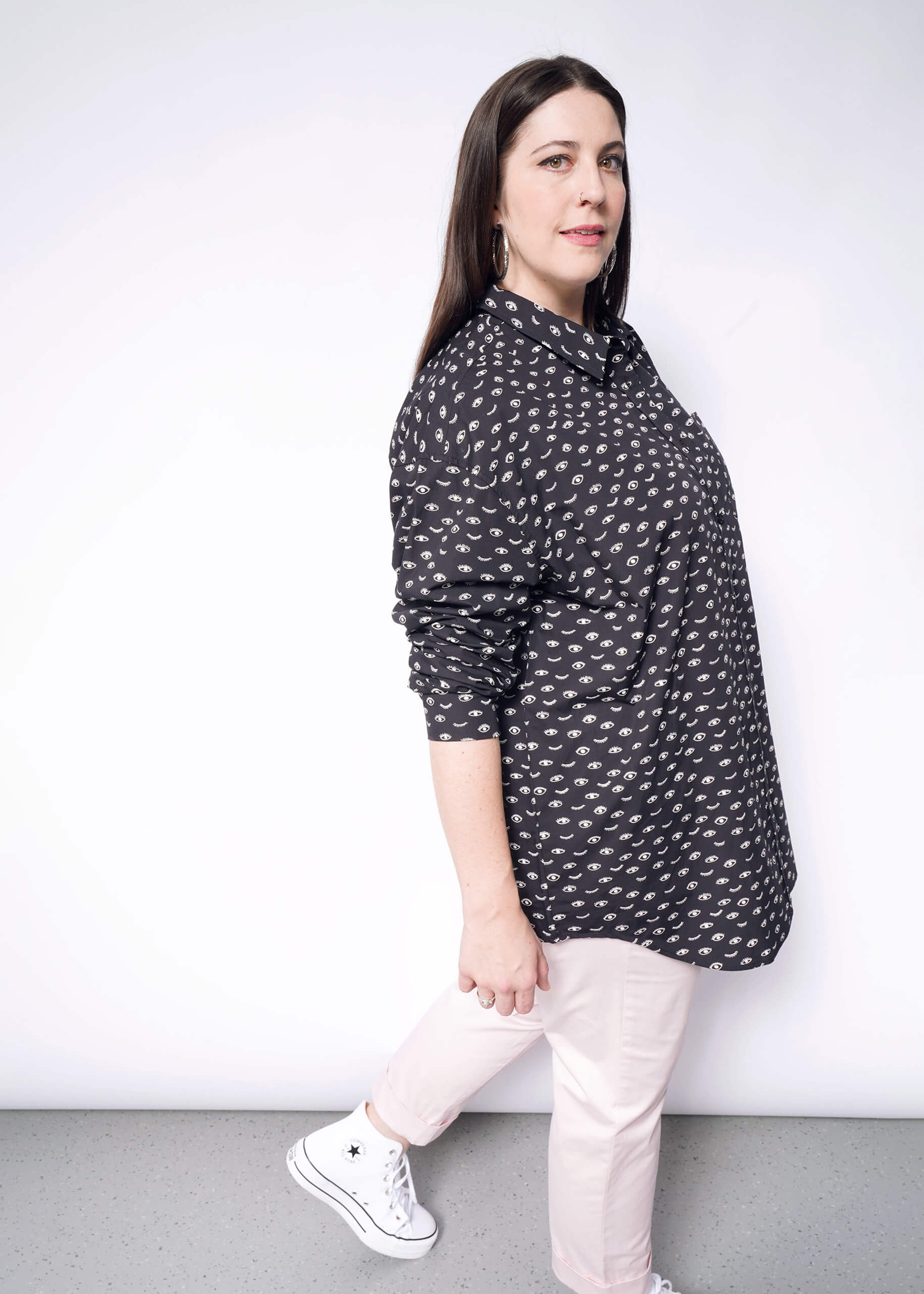 A person with long, dark hair stands against a white background wearing The Essential Long Sleeve Oversized Wrap Button Up. They pair it with light pink pants and white sneakers, gazing slightly to the side with a neutral expression.