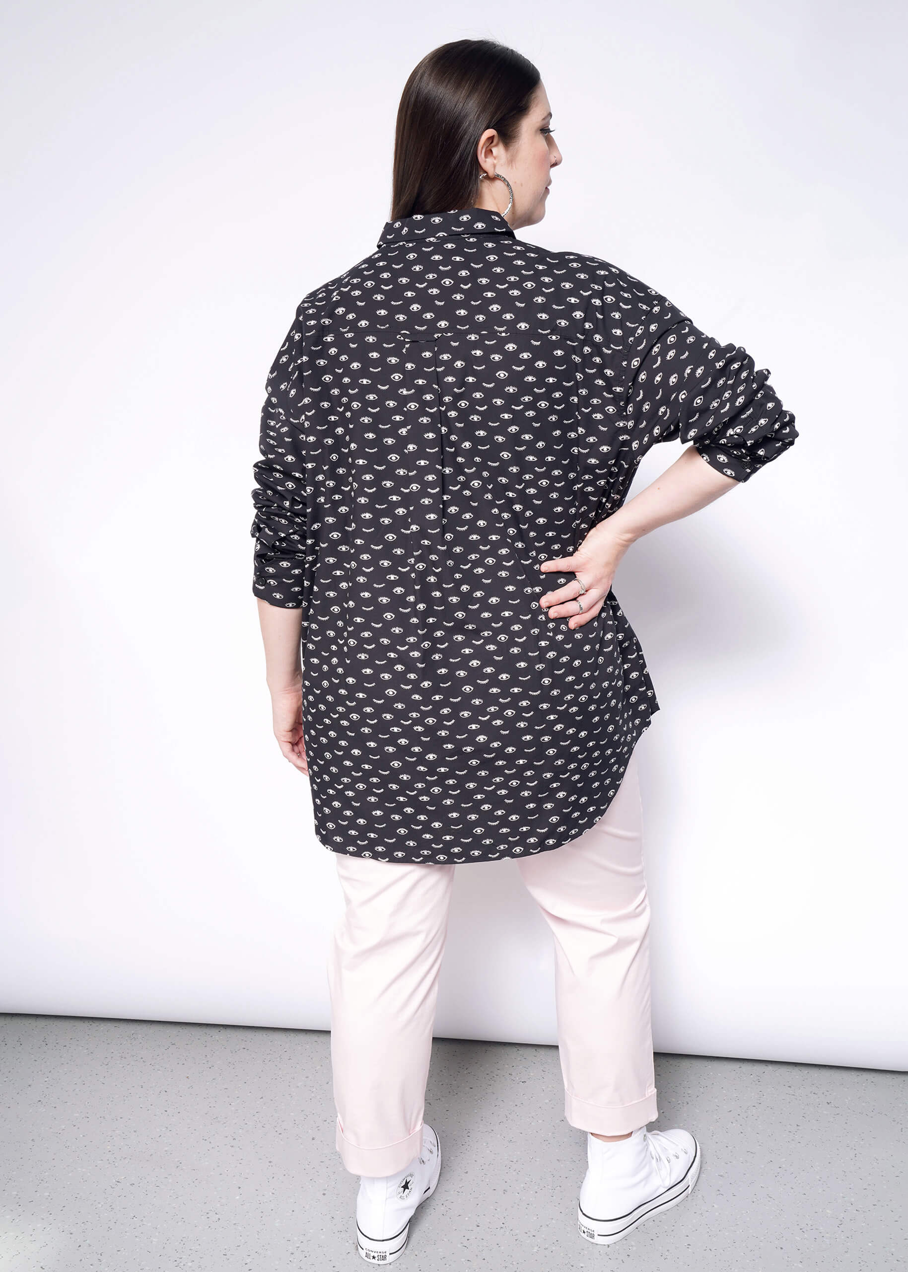 Rear view of a person with long dark hair wearing The Essential Long Sleeve Oversized Wrap Button Up in black with a white pattern, paired with light pink pants and white sneakers. They stand facing a white wall with one hand on their hip.