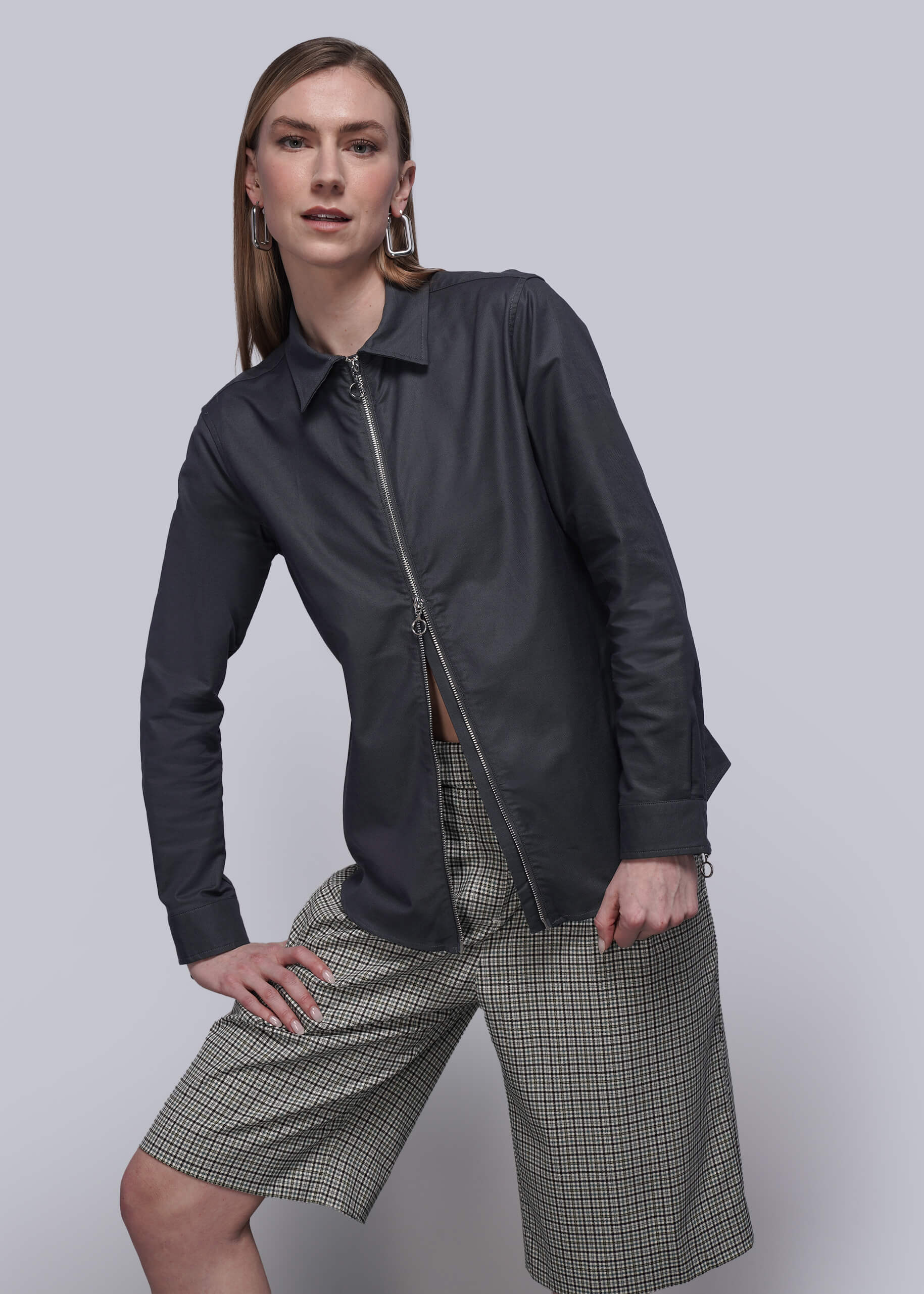 Person wearing a black zip up shirt, checkered shorts, and black boots on a plain background