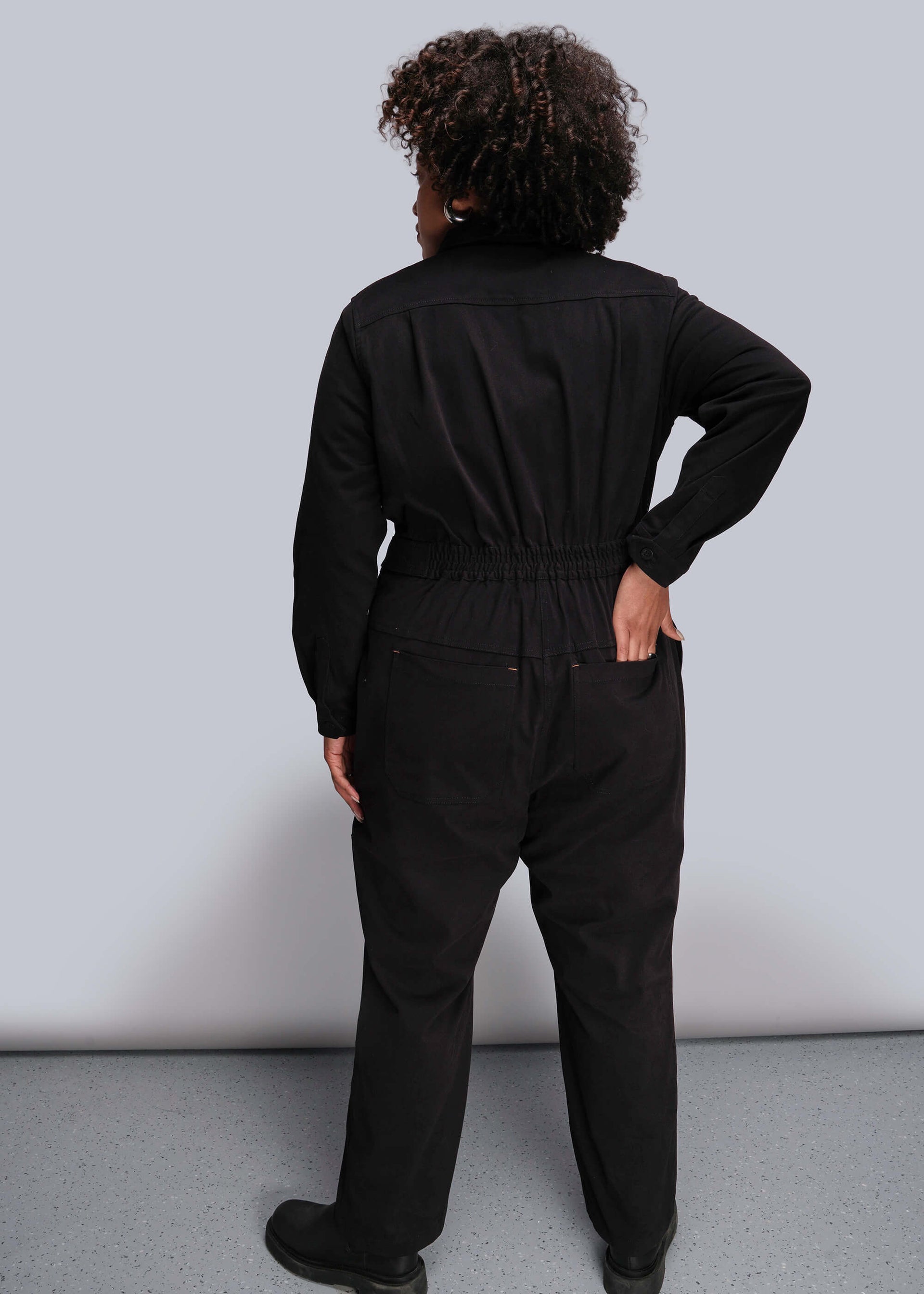1X=Person wearing a black jumpsuit standing against a plain background