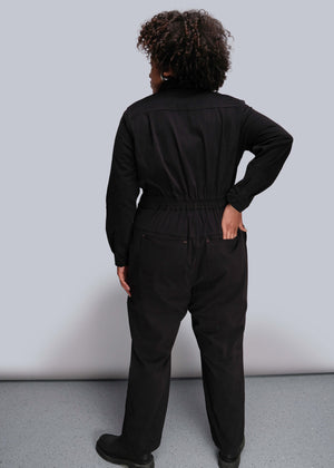 1X=Person wearing a black jumpsuit standing against a plain background