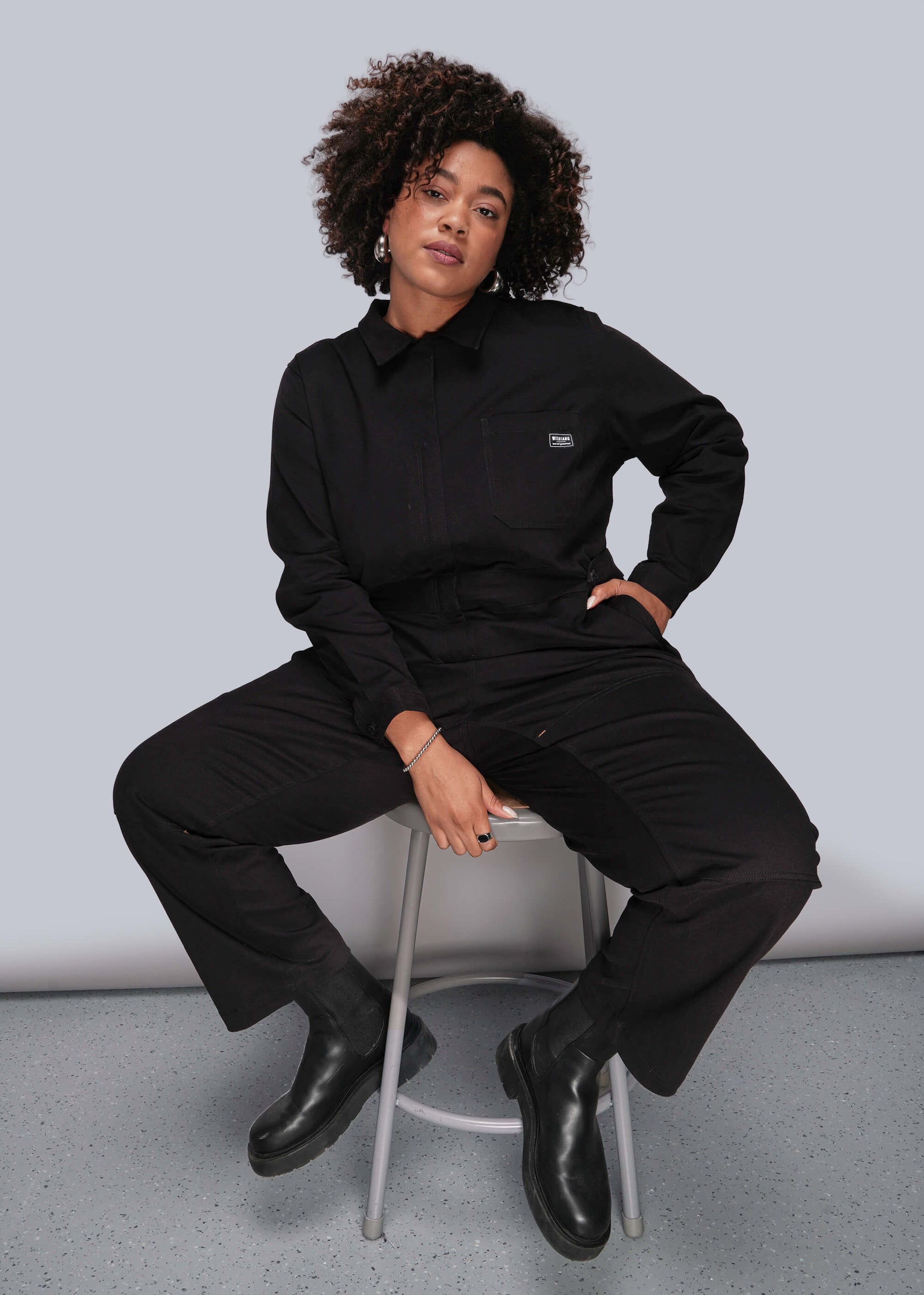 1X=Person wearing a black outfit sitting on a chair against a plain background