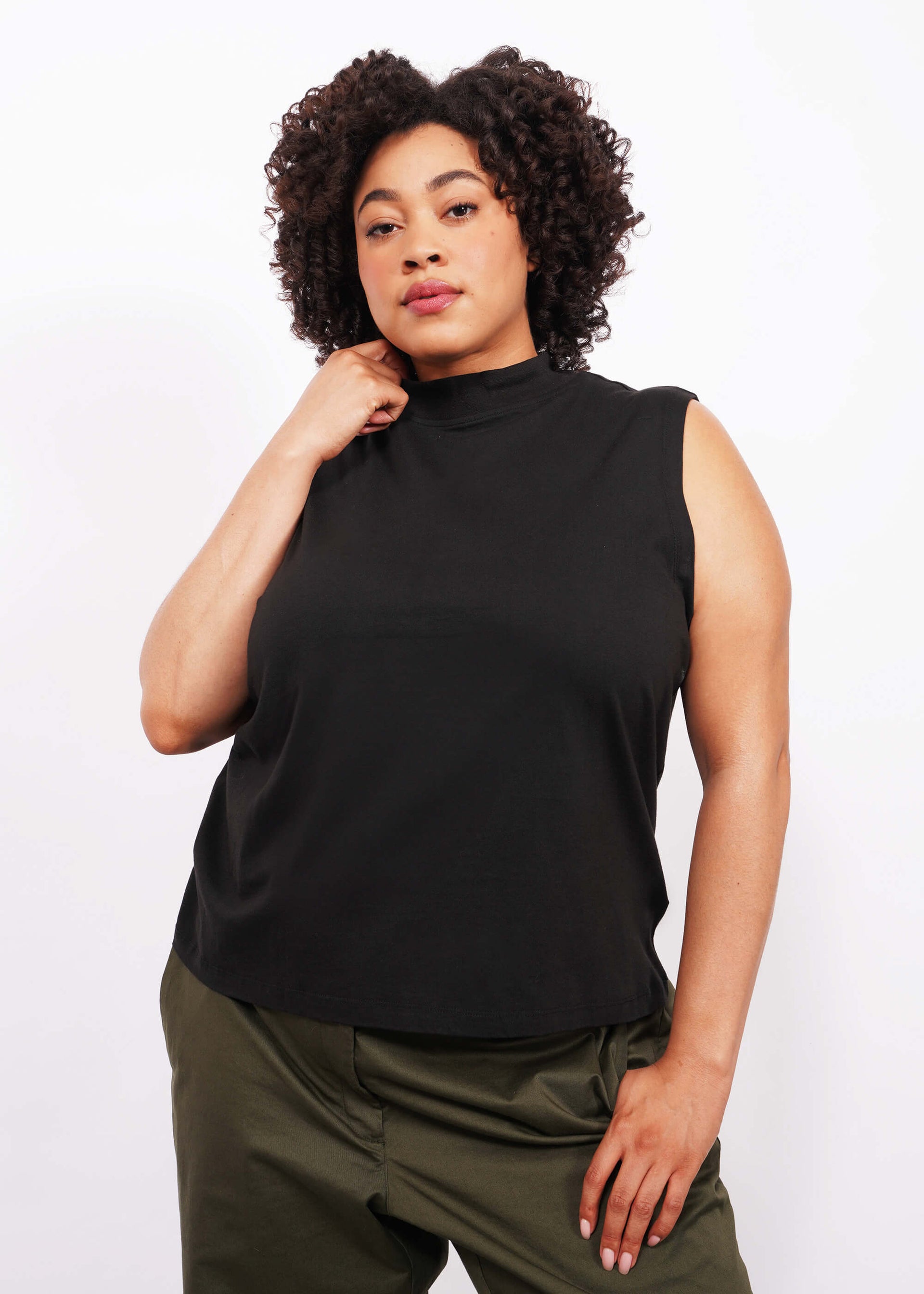 A person with short curly dark hair stands against a white background, wearing The Essential Mock Neck Tank and dark green pants, with one hand touching the shirt's collar while the other is in the pant pocket, and looking directly at the camera.